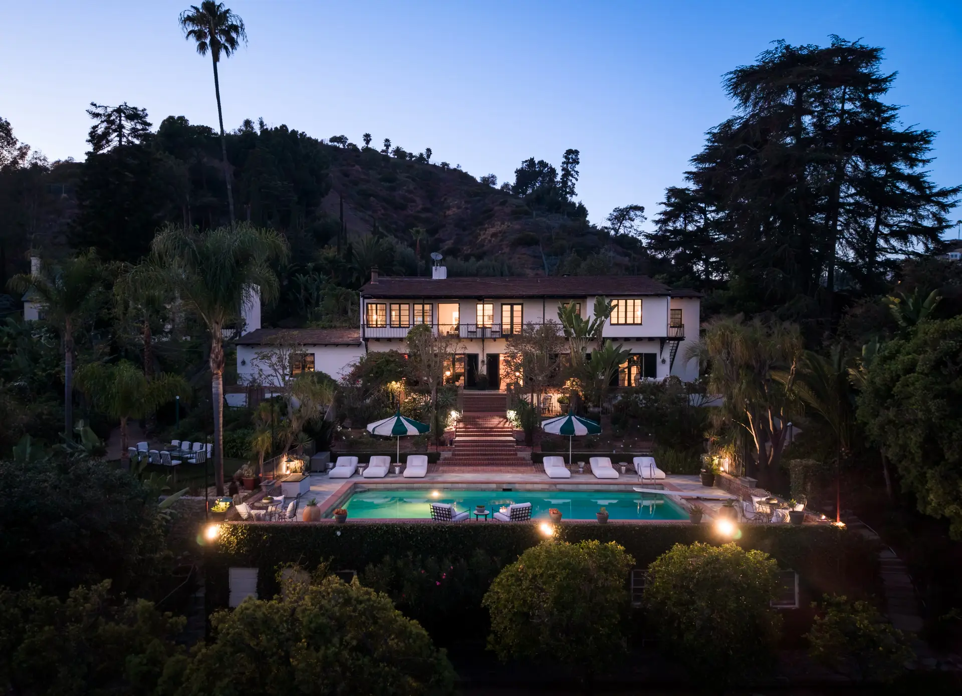 Illuminated hillside villa with a swimming pool and palm-filled garden at dusk.