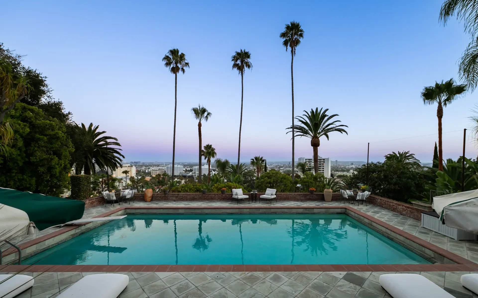 Rectangular outdoor swimming pool bordered by lounge chairs and tall palm trees overlooking a distant city skyline at dusk.