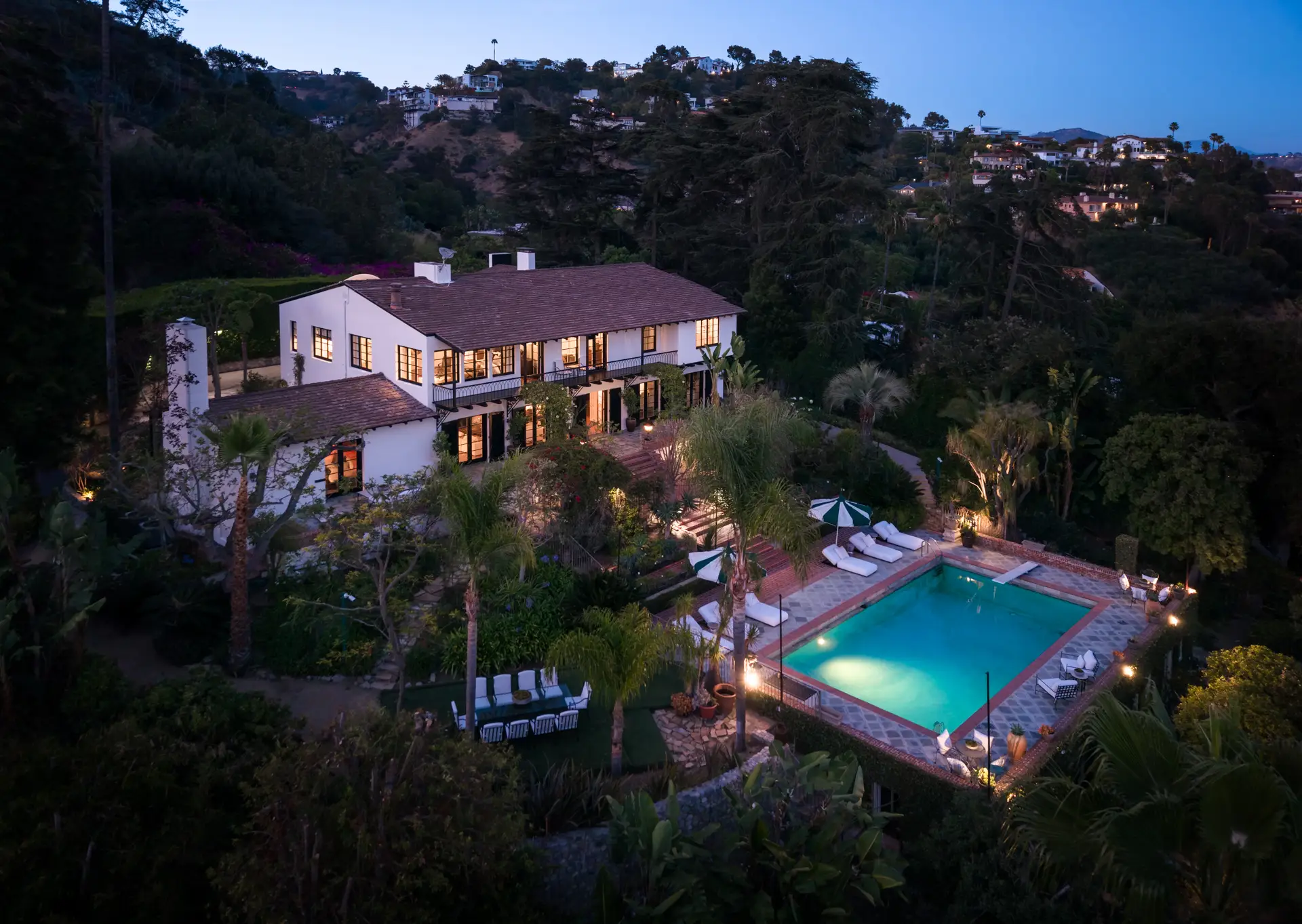 Large illuminated hillside home with landscaped gardens and a swimming pool at dusk.