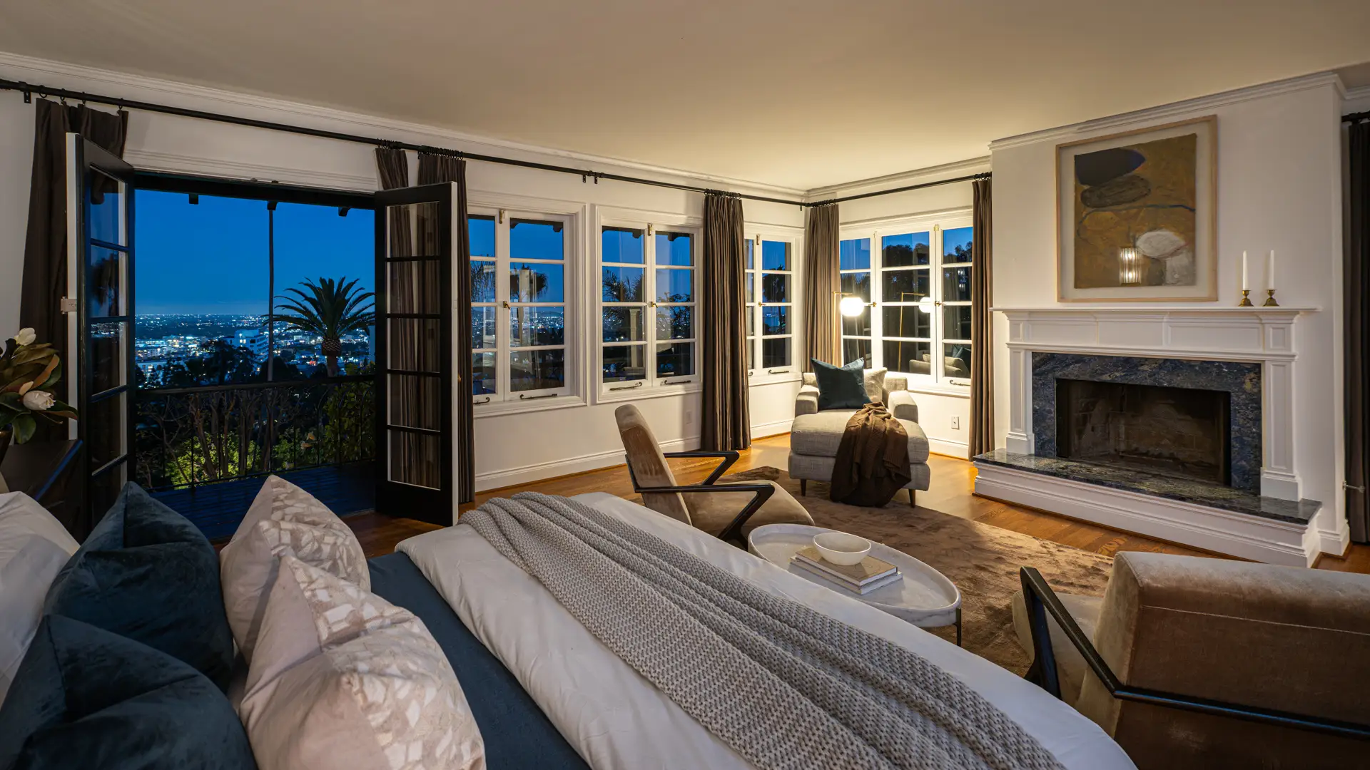 Luxury bedroom with open balcony doors, city lights view, fireplace, seating area, and large windows at dusk.