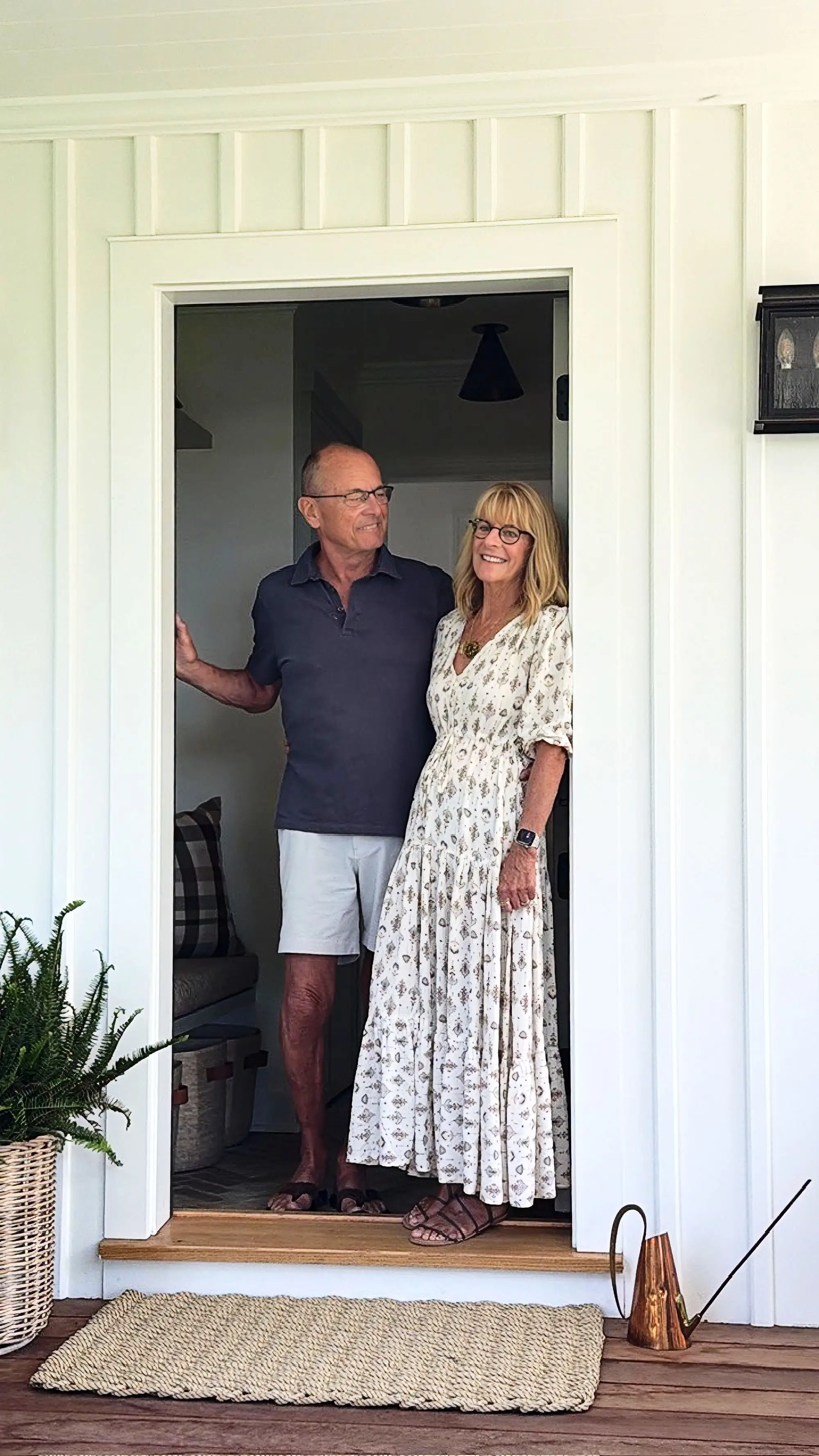 Two people stand smiling in a white doorway on a porch with a woven mat and potted fern.