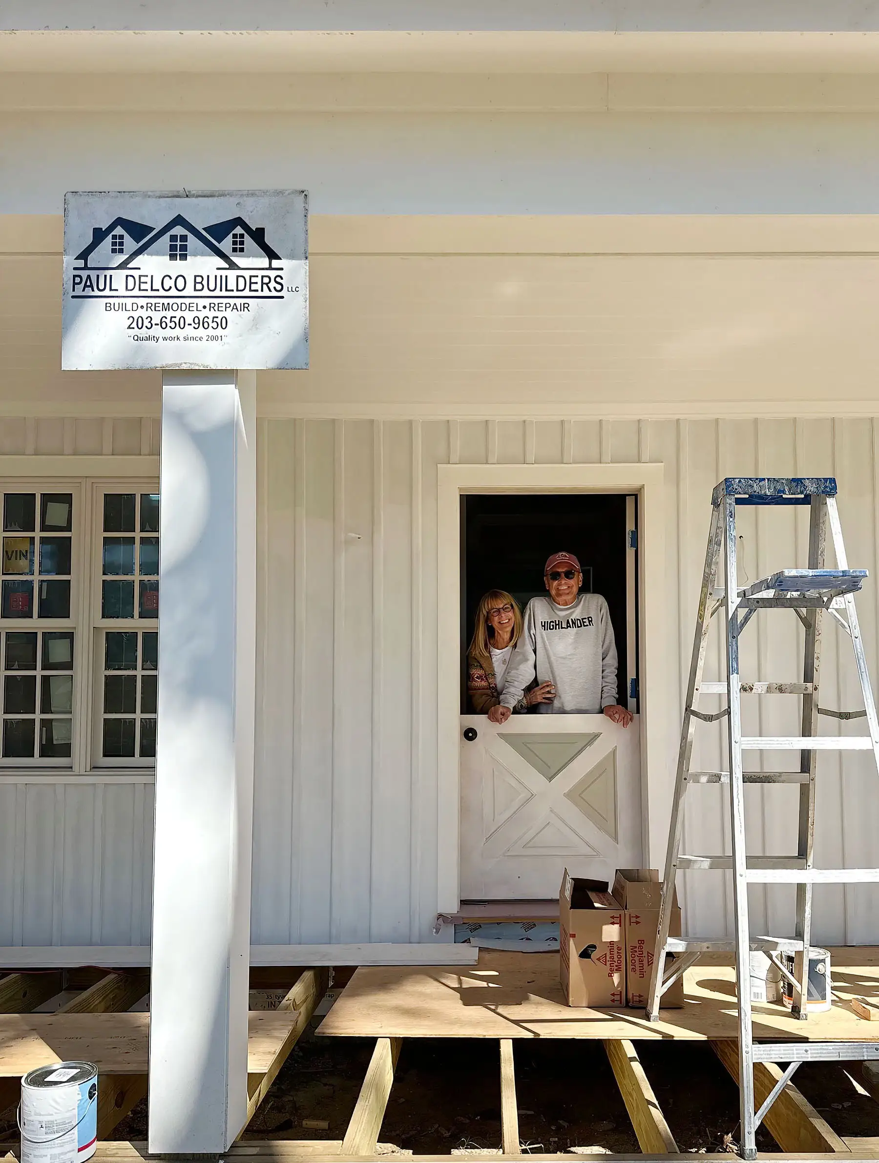 Two people stand in a half door during porch construction beside a ladder and Paul Delco Builders LLC sign.