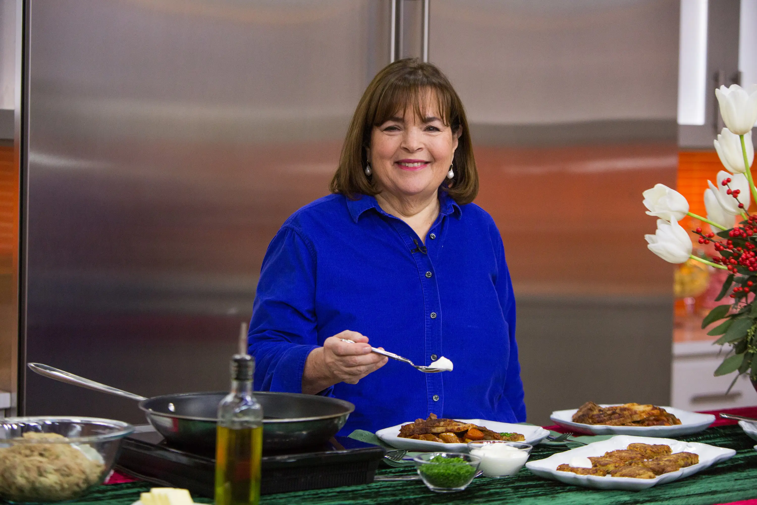 Ina Garten stands at a kitchen counter with a skillet, olive oil, plated food, and white tulips.