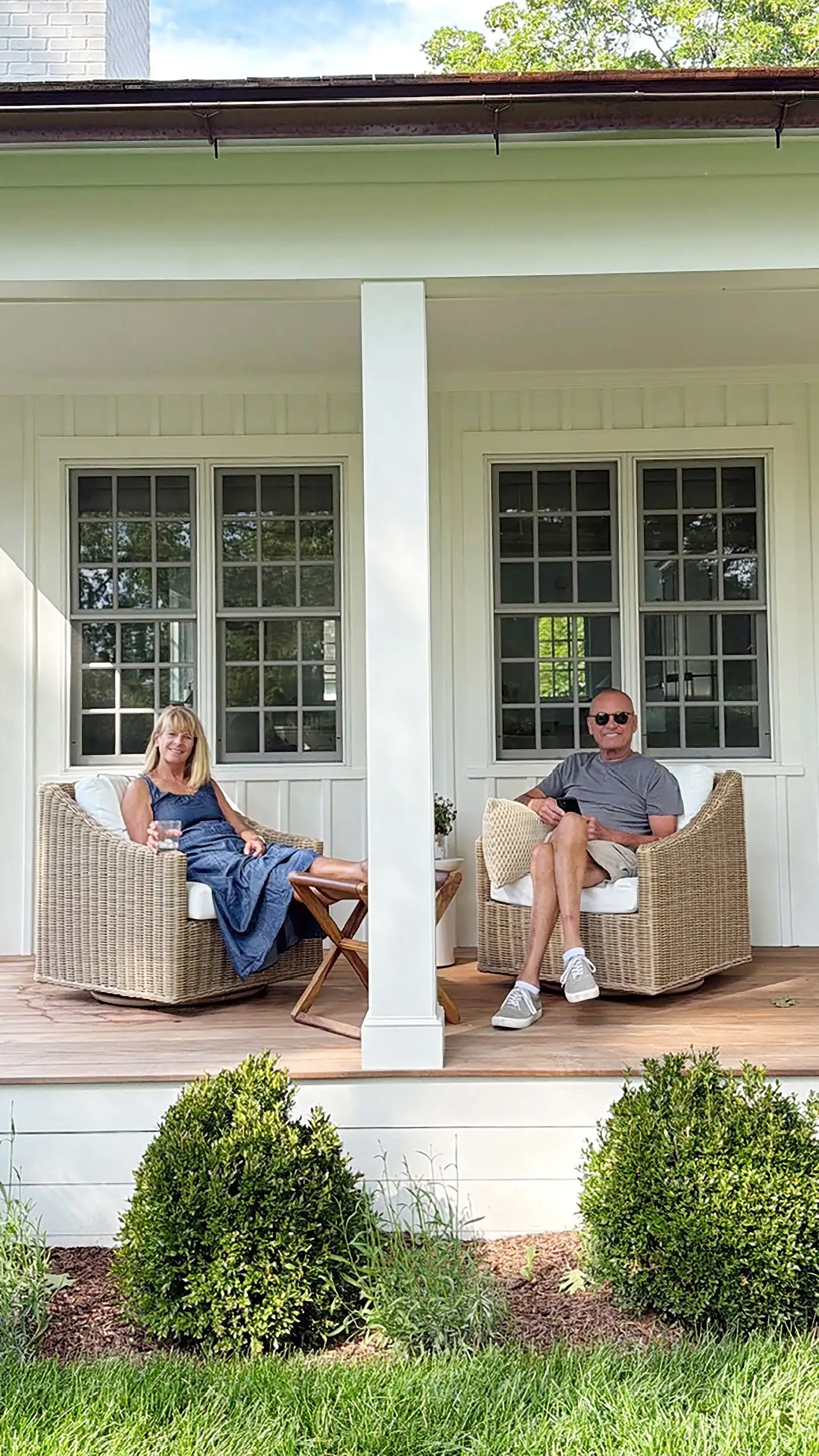 Two adults relax in wicker chairs on a covered white porch with shrubs and lawn in front.