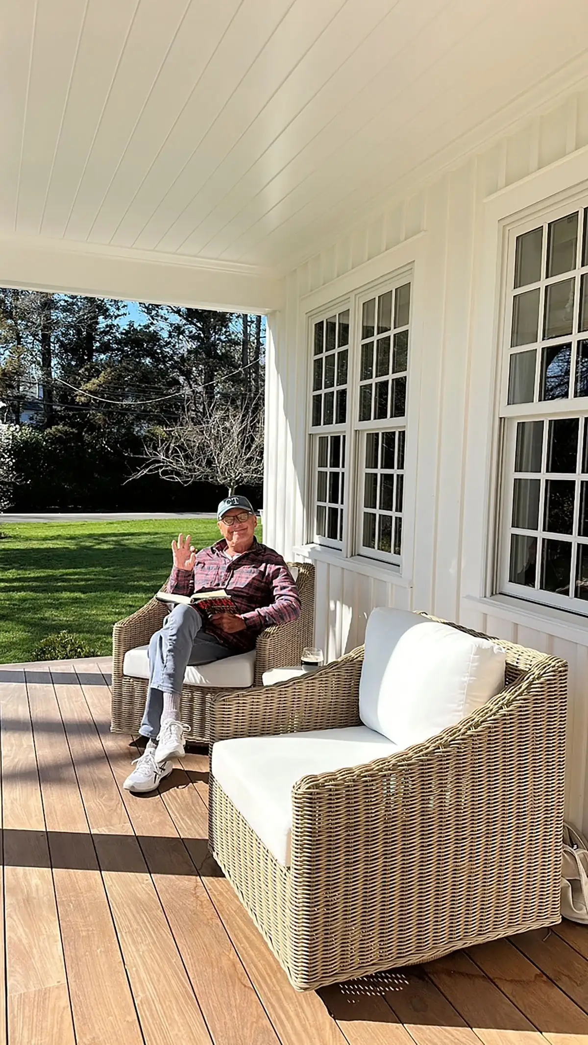 A person waves while reading on a wicker chair on a sunny porch beside large windows.