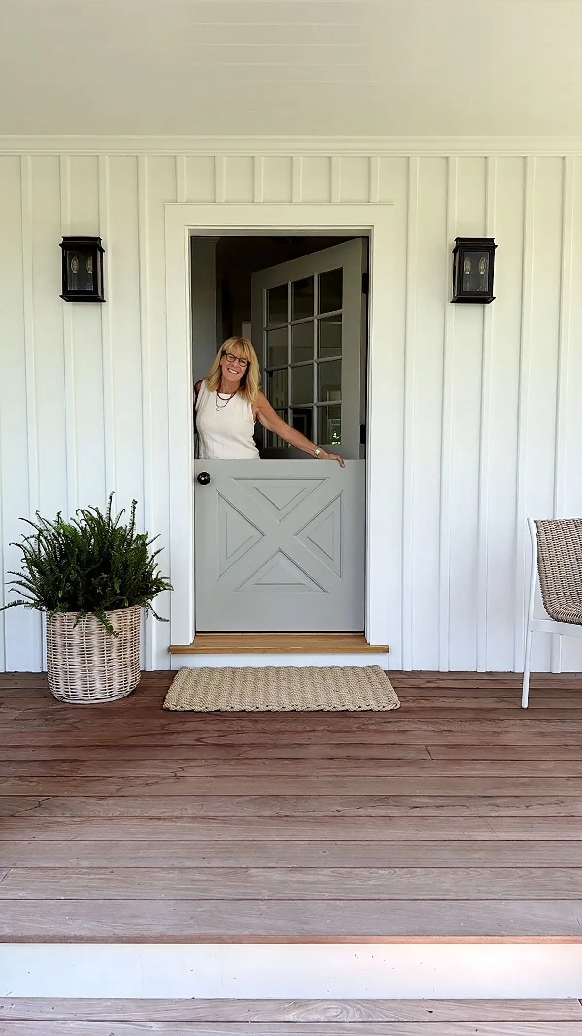 A smiling person stands behind a Dutch door on a covered porch with wicker furniture and potted greenery.