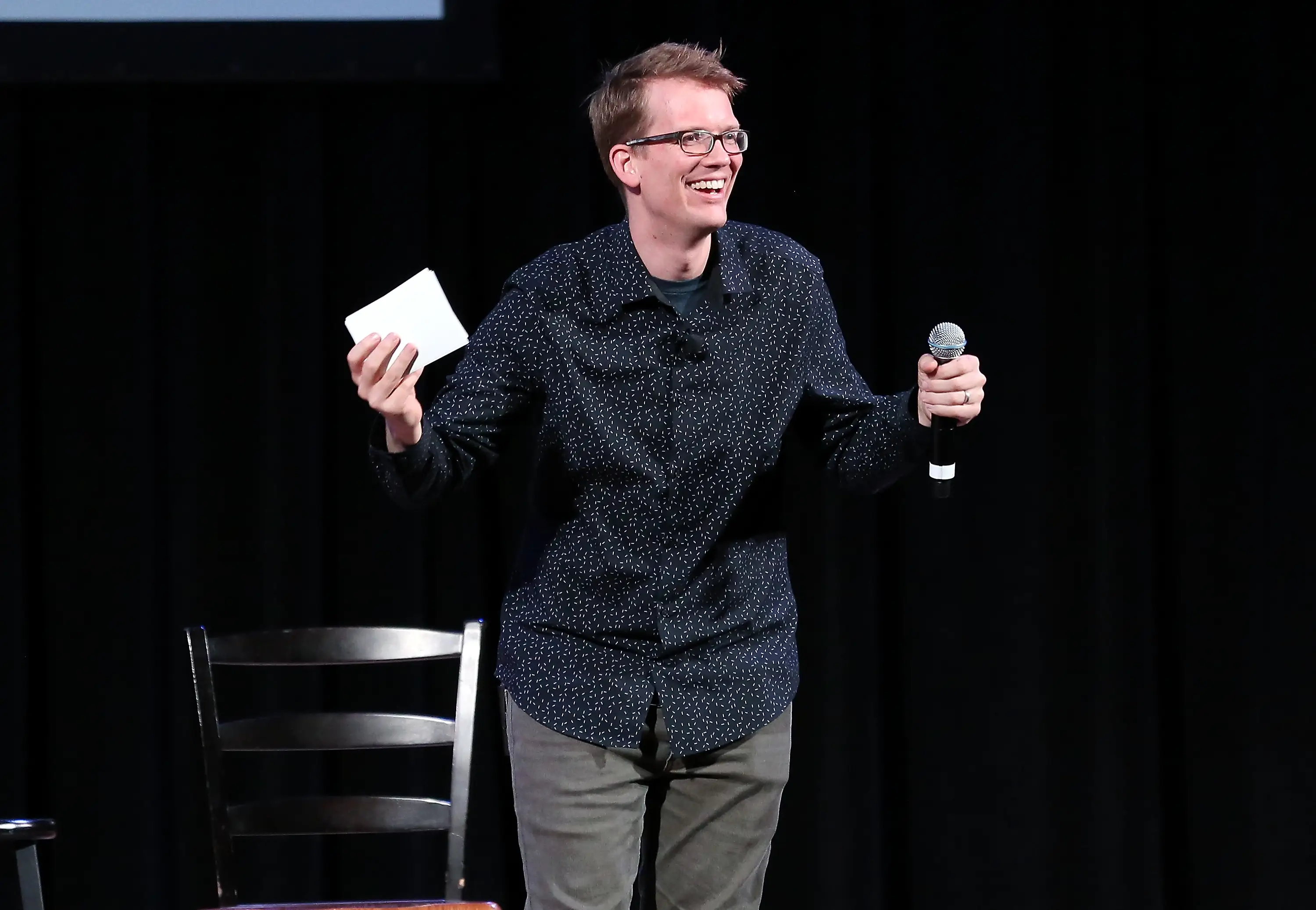 Hank Green smiles onstage while holding a microphone and a note card beside a chair.