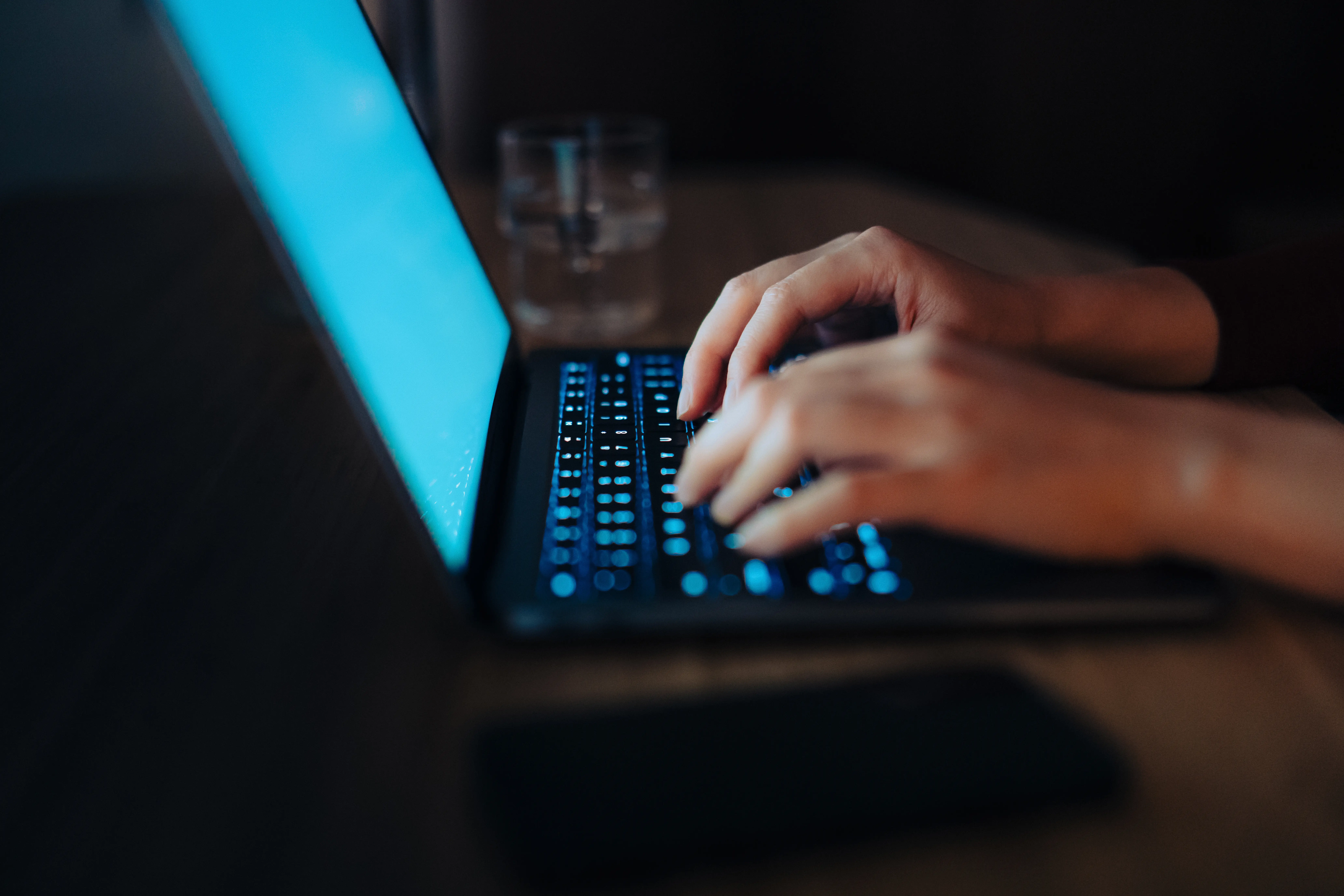 Hands type on a glowing laptop keyboard in a dimly lit workspace with a blurred phone nearby.