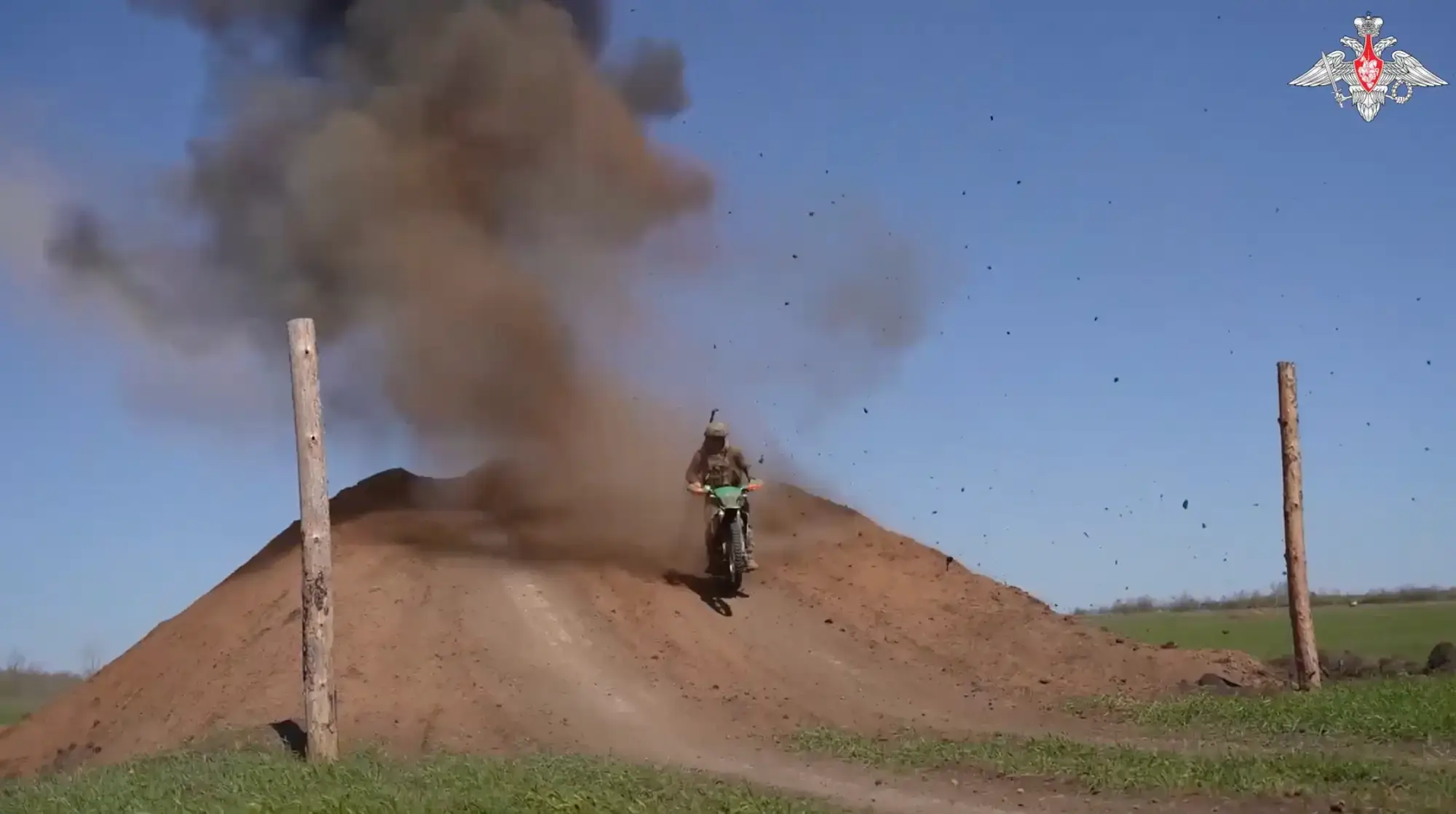 A motorcyclist rides over a dirt mound as a blast throws smoke and debris behind them.