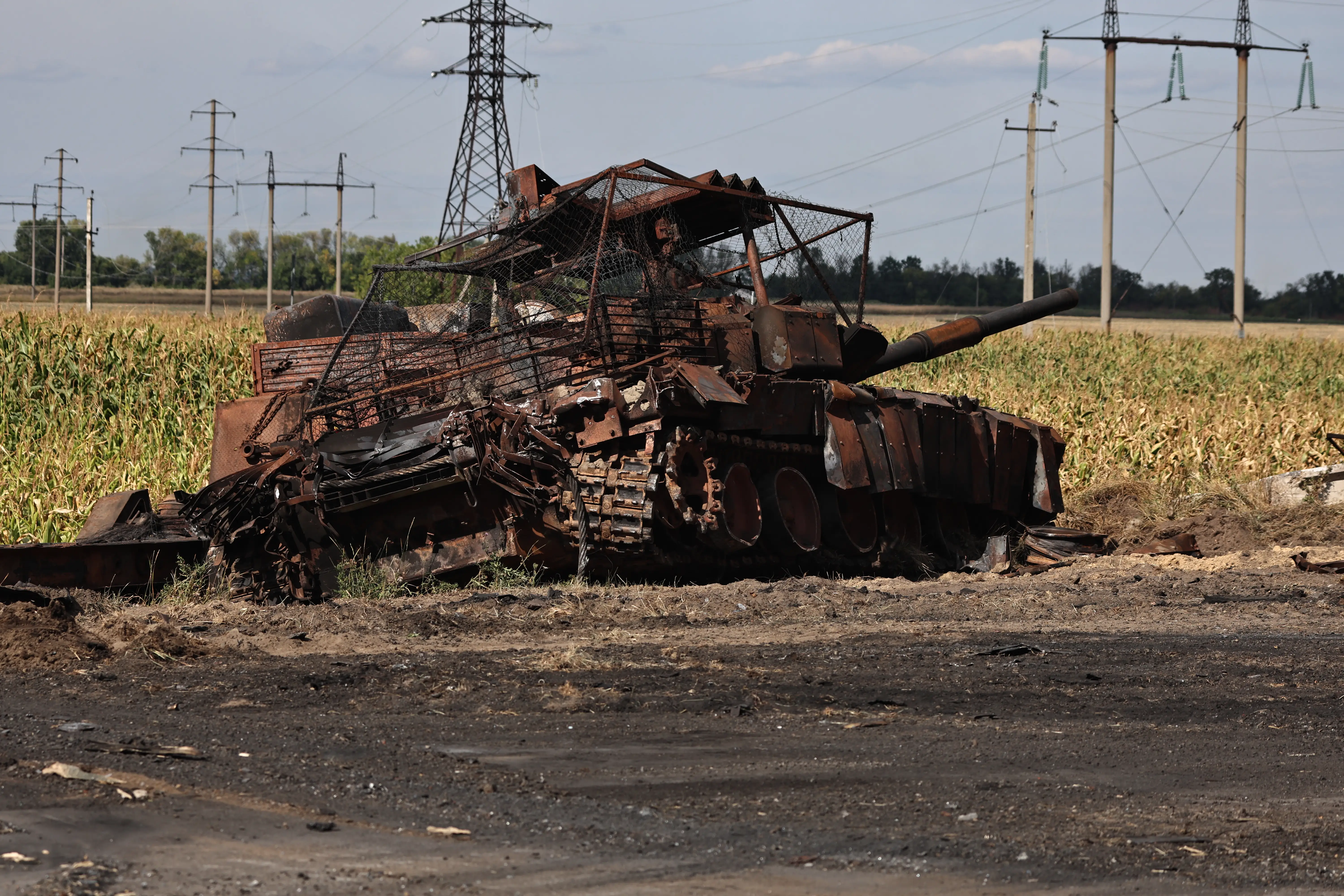 Burned-out tank wreckage sits beside a crop field with power lines in the background.