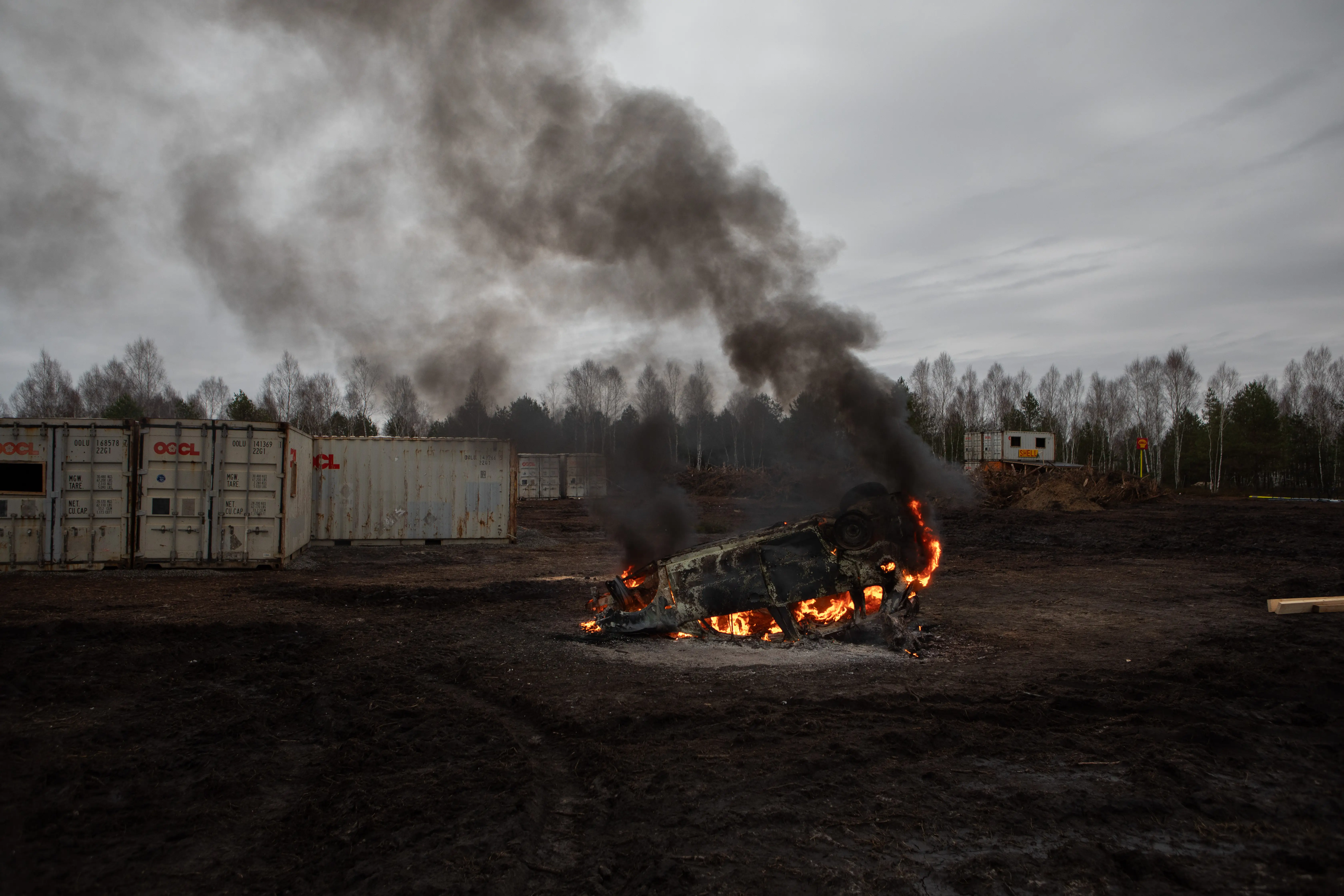 Overturned vehicle burns in a muddy clearing beside shipping containers as thick black smoke rises near a tree line.