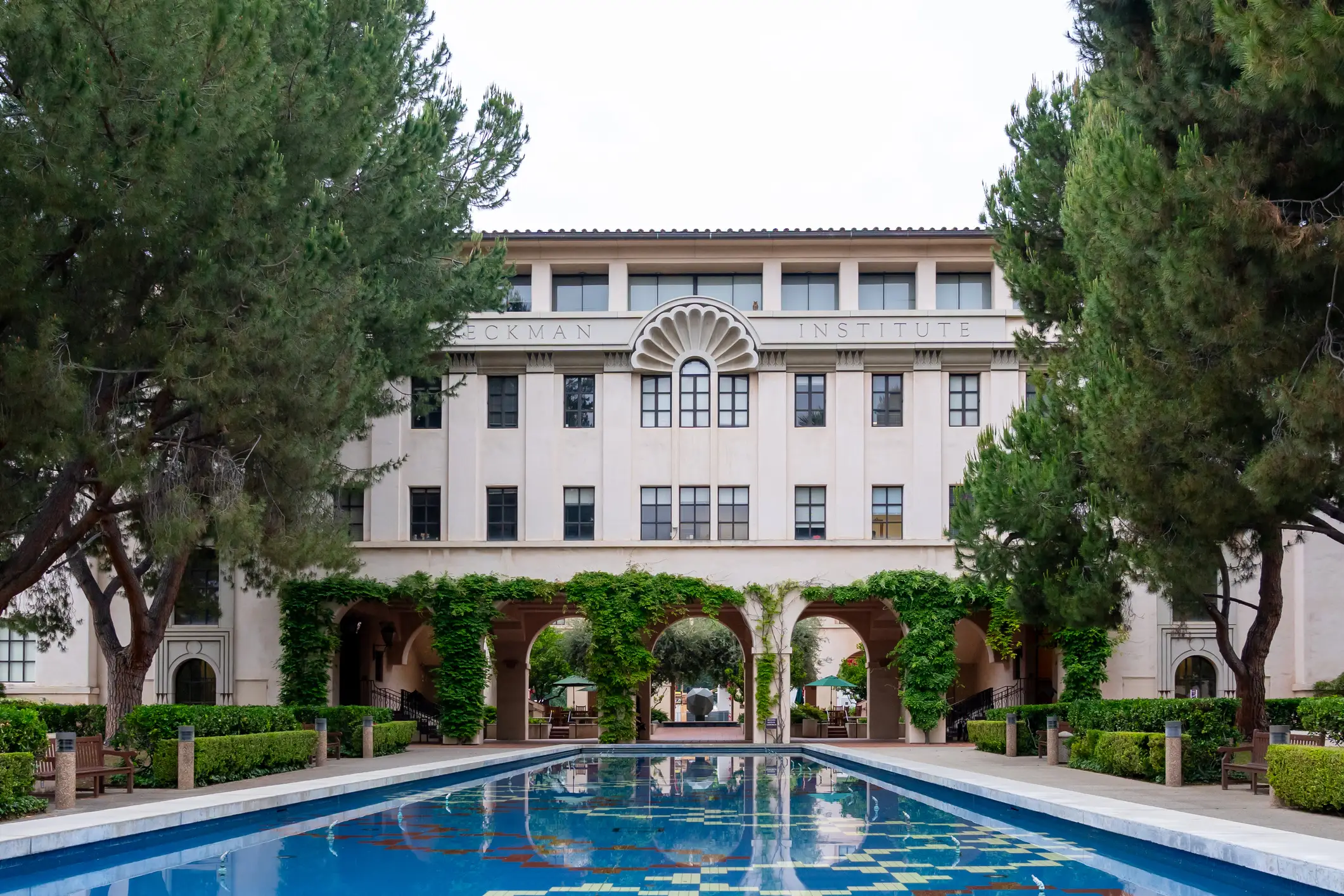 Caltech Beckman Institute building framed by trees with an arched walkway and reflecting pool in front.