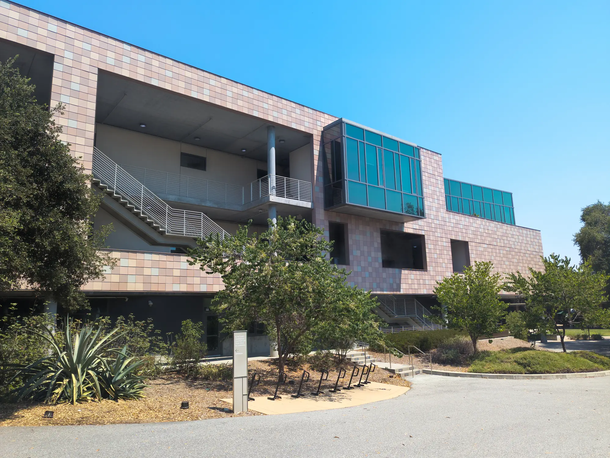 Modern tiled building with blue glass windows, exterior stairs, trees, and bicycle racks under a clear sky.