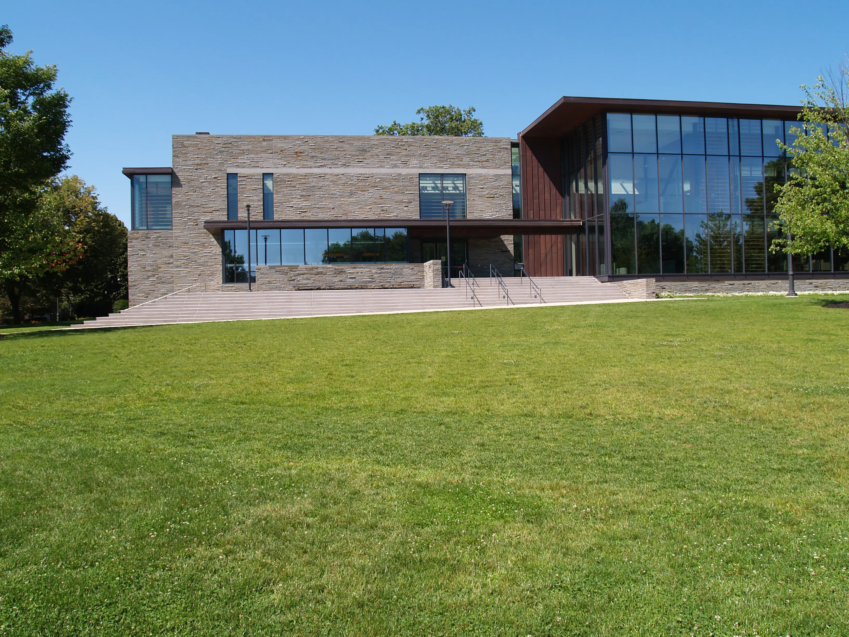 Modern stone-and-glass Skillman Library at Lafayette College with broad steps sits behind a large green lawn under a clear blue sky.