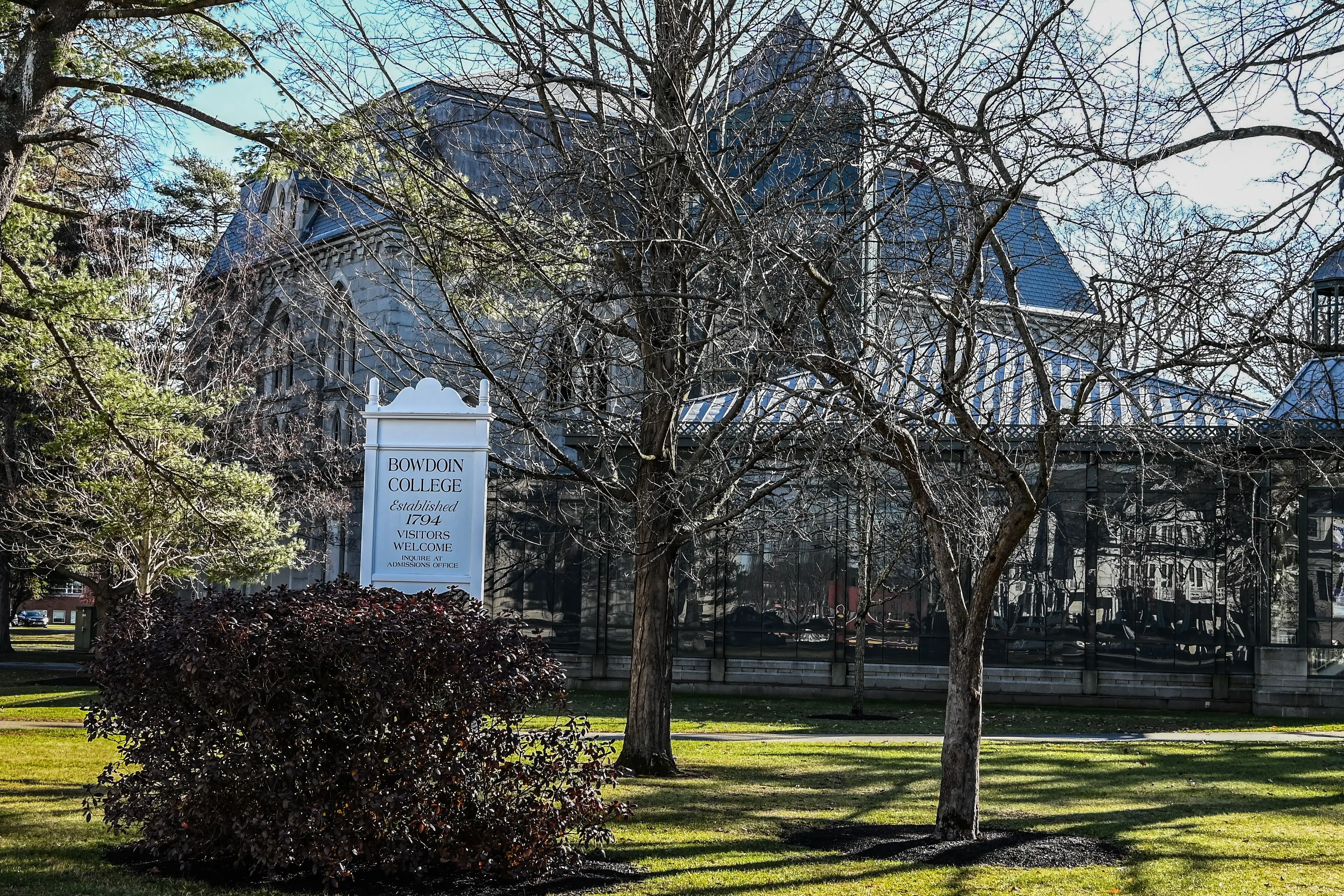 Bowdoin College sign stands on a tree-lined lawn in front of campus buildings.