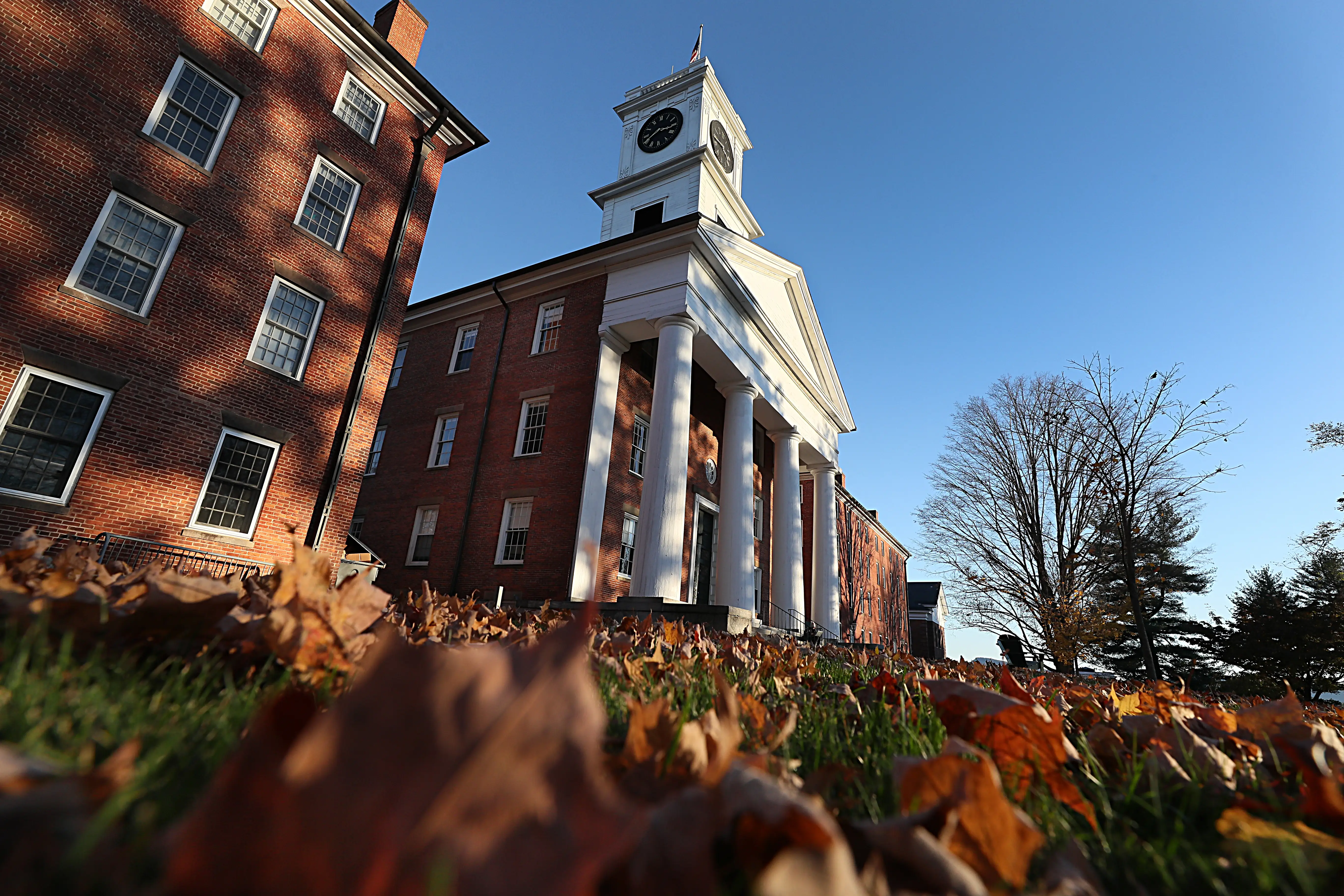 Amherst College's red brick building with white columns and a clock tower rises behind autumn leaves under a clear blue sky.