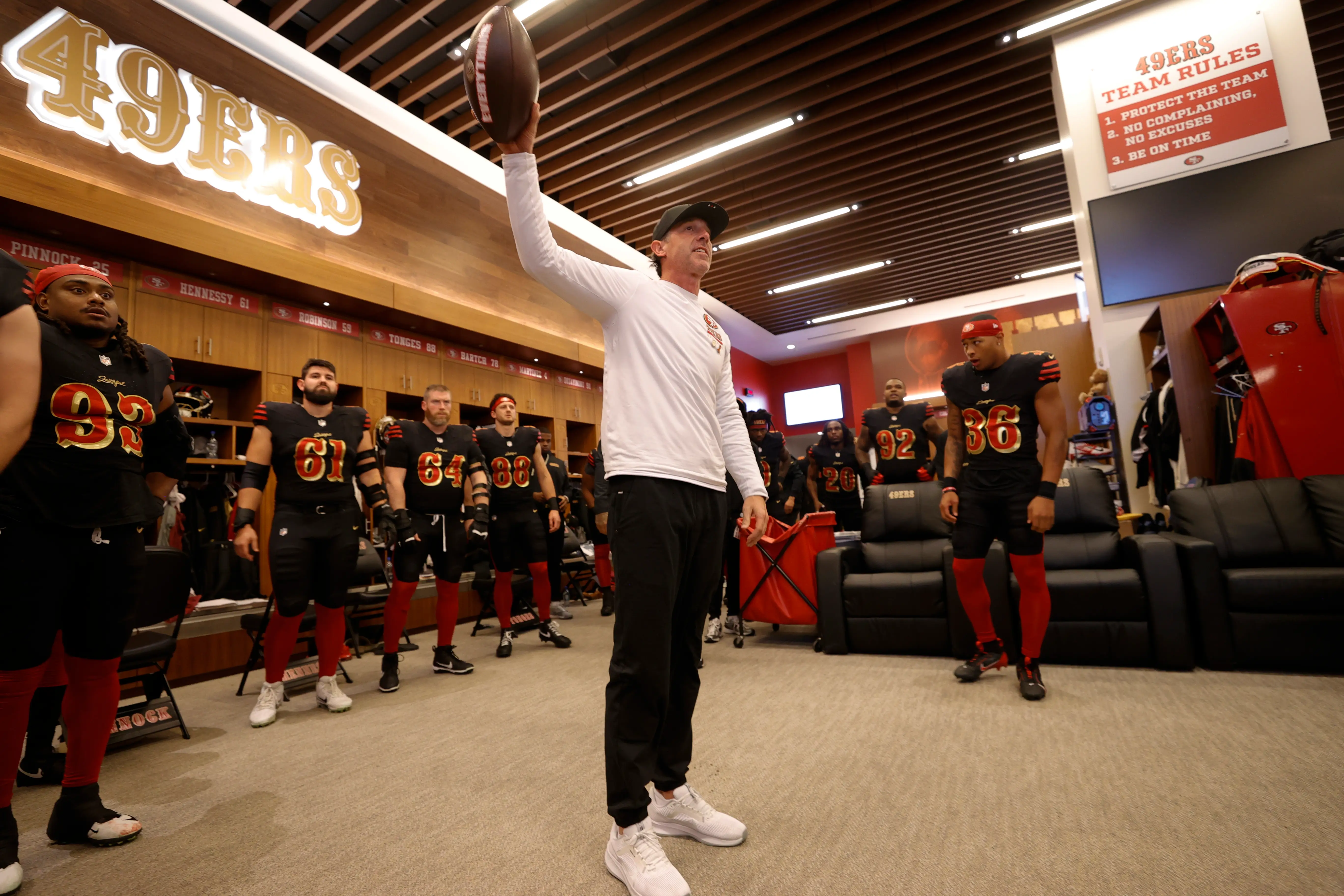 San Francisco 49ers head coach Kyle Shanahan holds a football in his right hand while speaking to his team in a locker room.