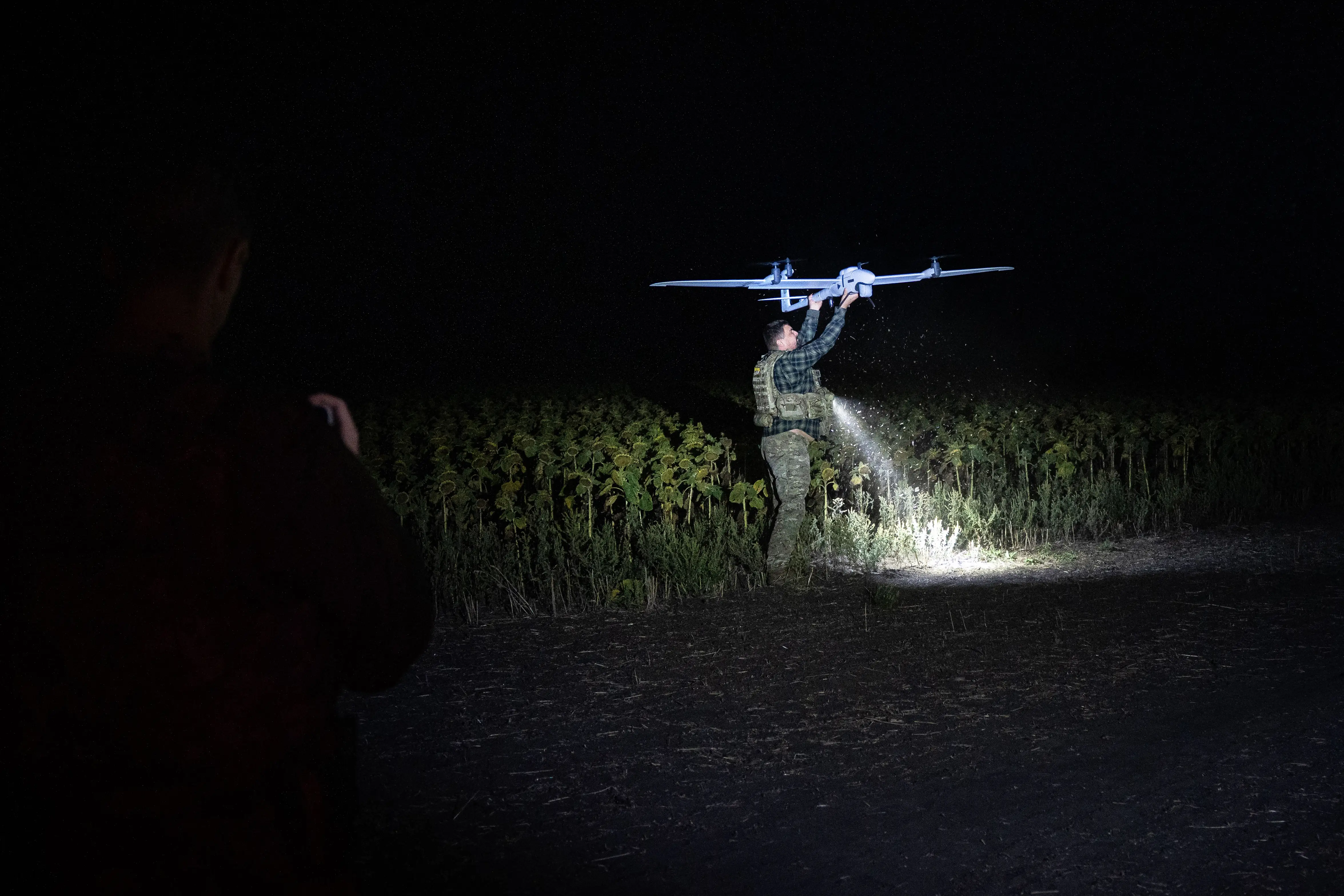 A person lifts a large drone above a crop field at night while illuminated by a bright light.