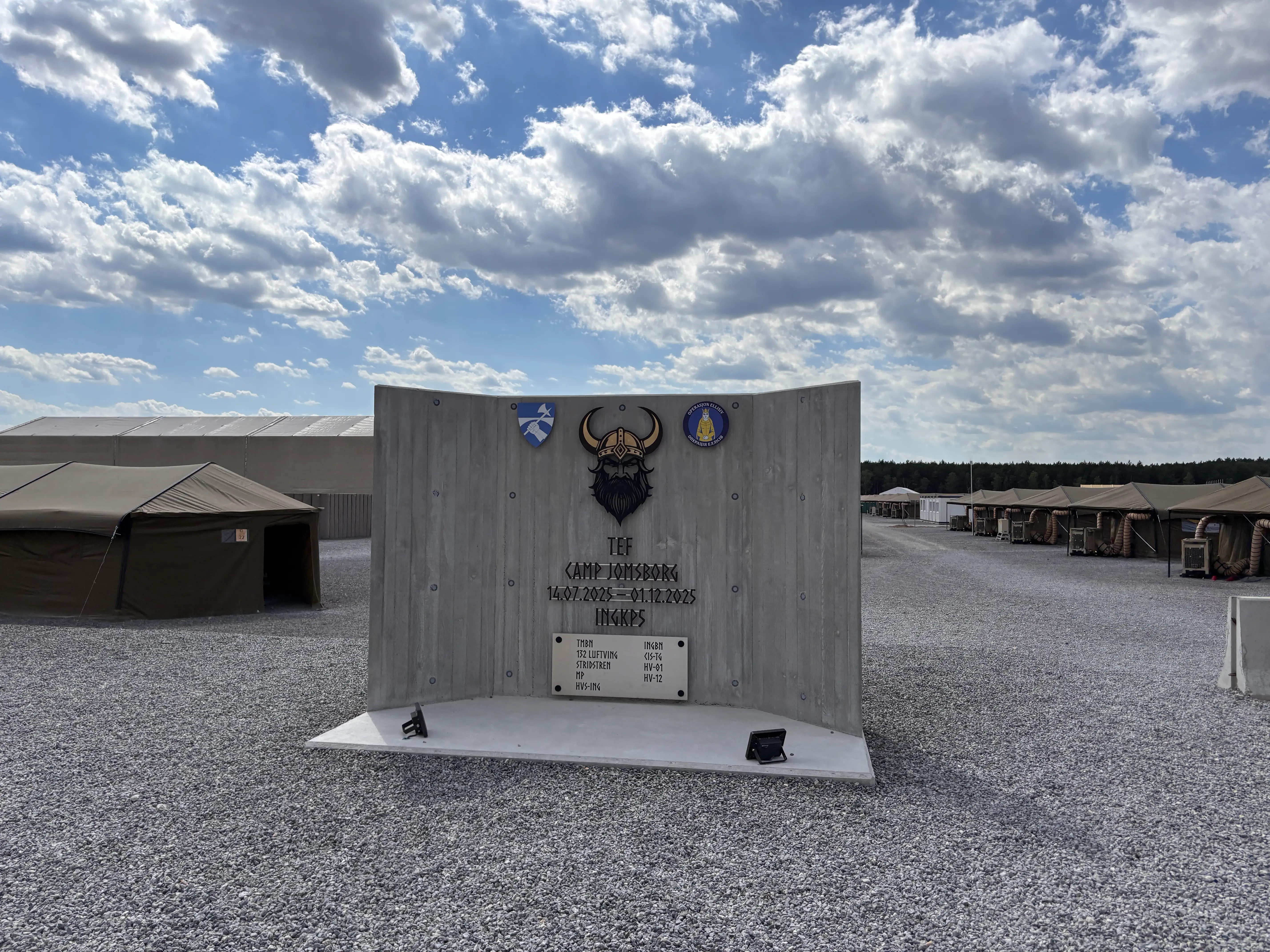Concrete camp sign with a Viking emblem stands between rows of tents on a gravel site under a cloudy sky.
