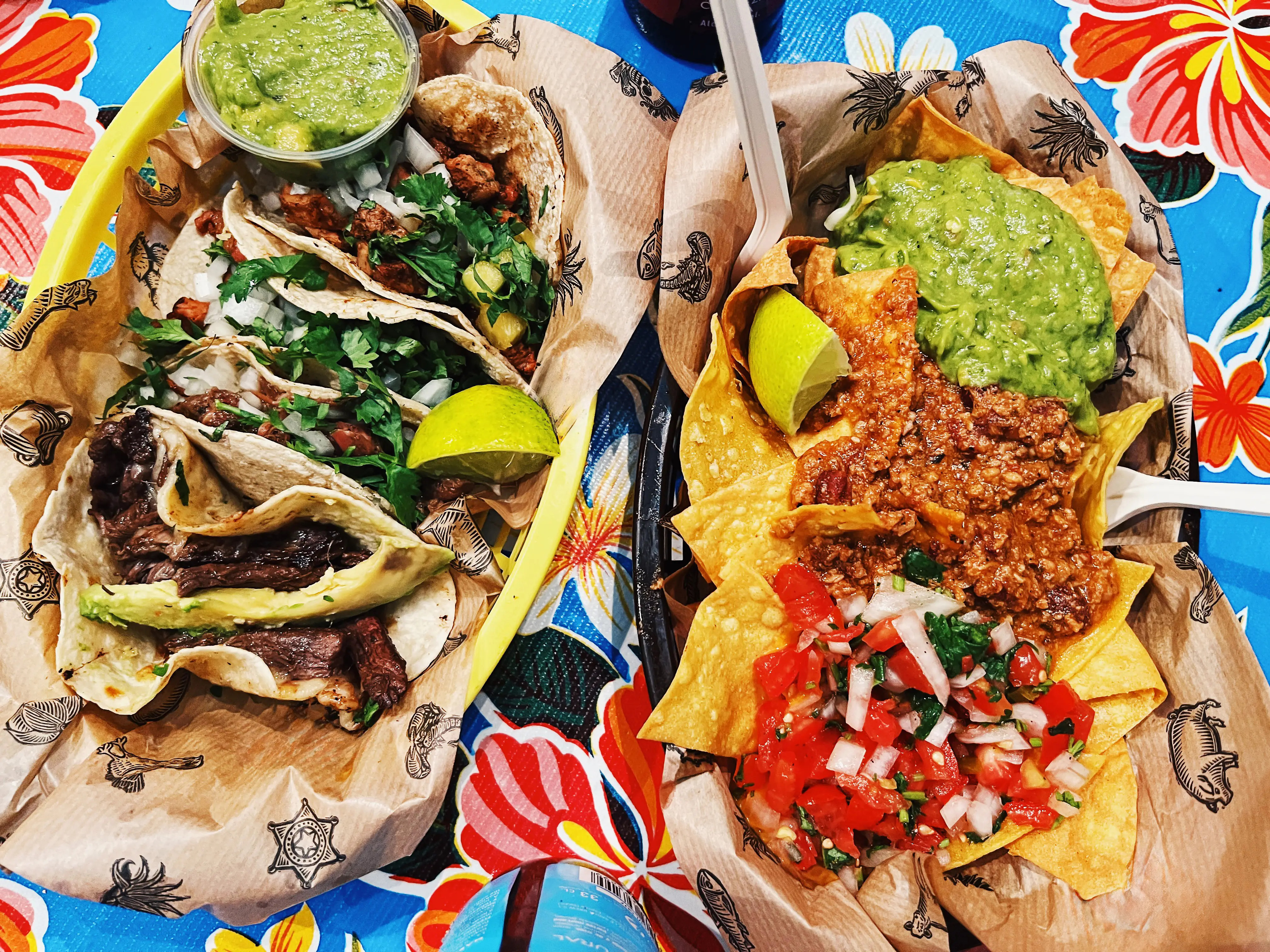 Tacos and nachos with guacamole, salsa, lime wedges, and herbs sit on patterned paper plates over a colorful floral tablecloth.