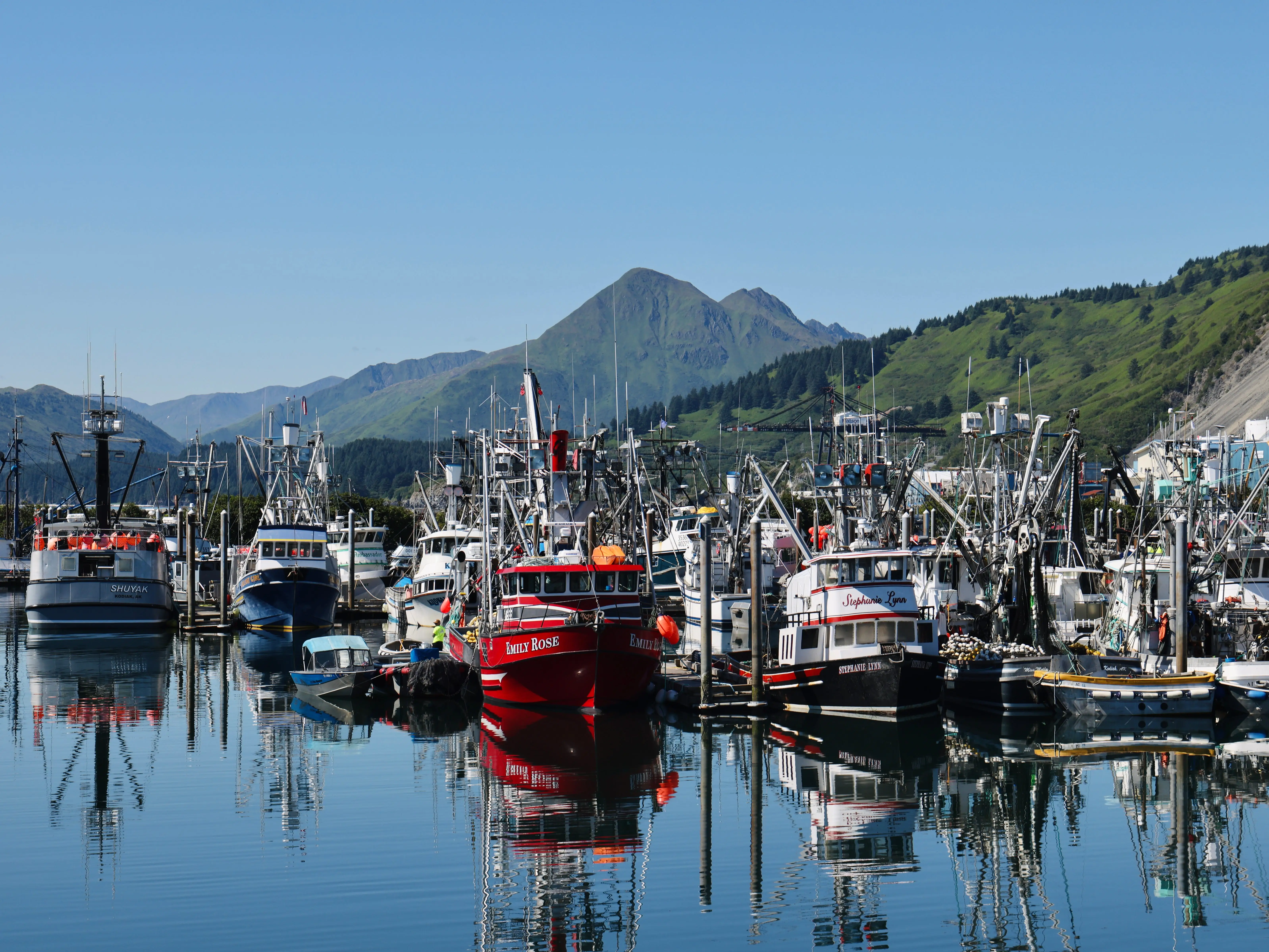 Fishing boats crowd a calm harbor with mountain slopes reflected in the water near a remote island off the coast of Alaska.