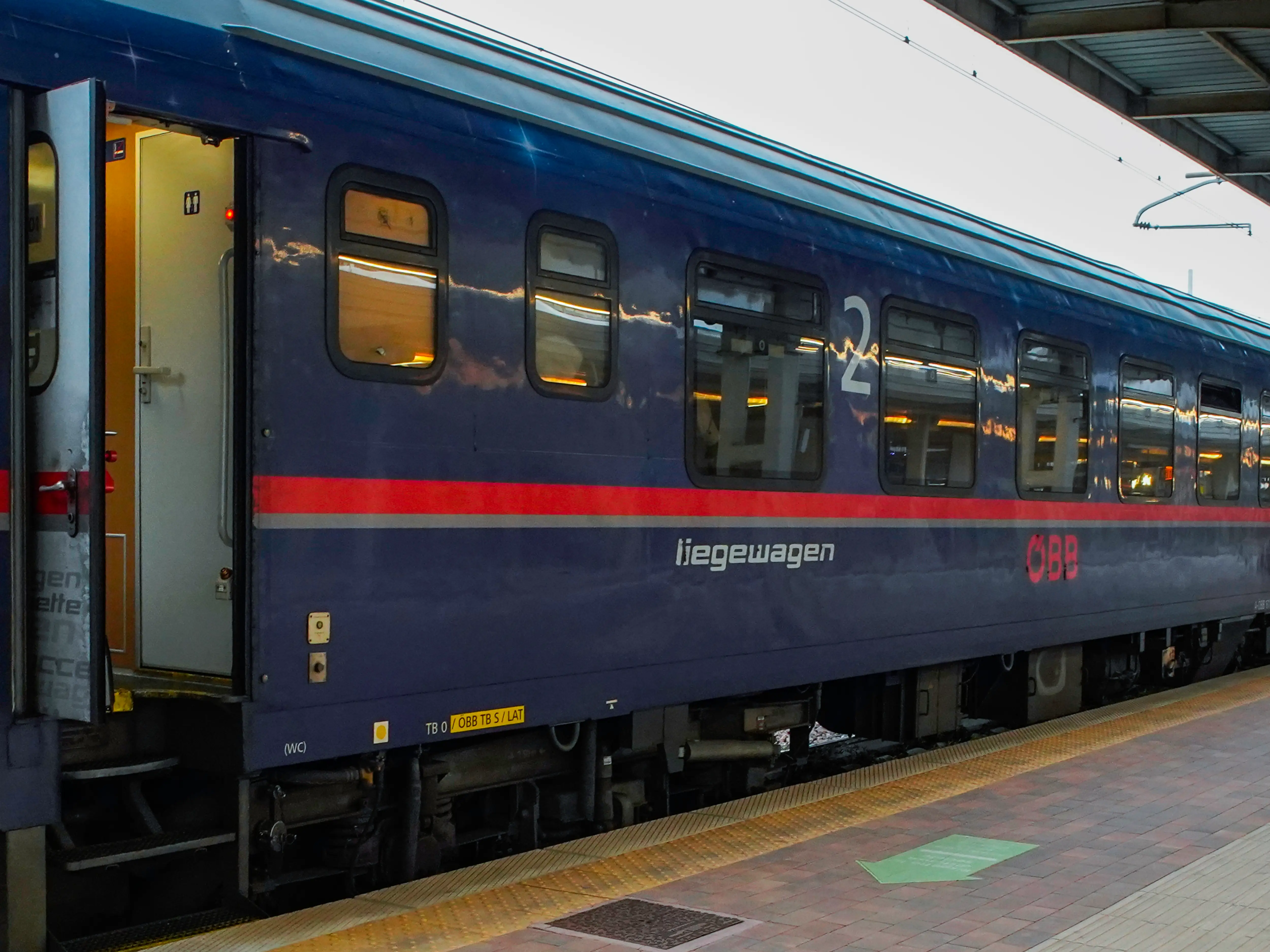 Blue ÖBB second-class sleeper coach stands at a railway platform with an open door.