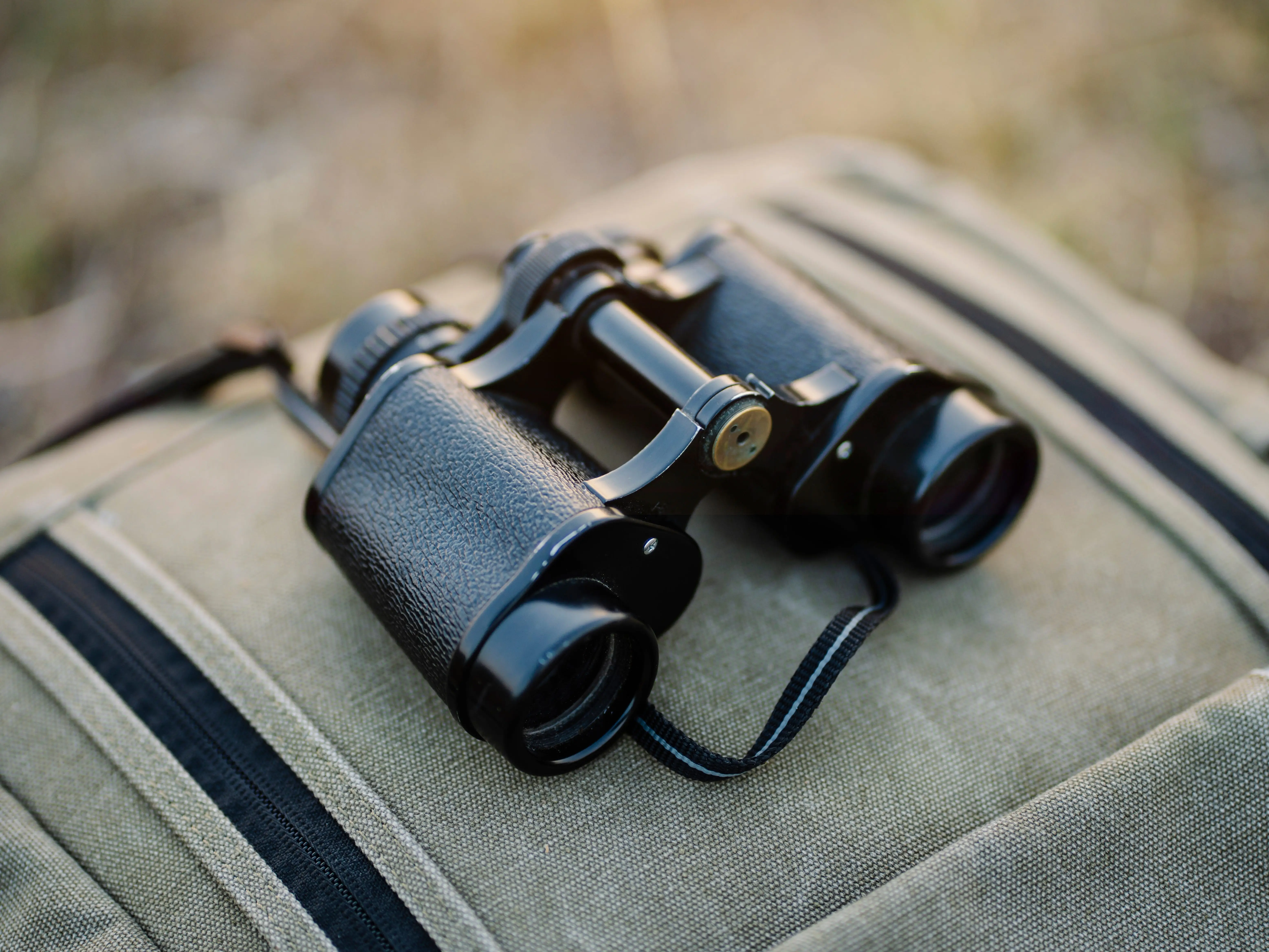 Black binoculars rest on a green canvas backpack in an outdoor setting with a blurred background.