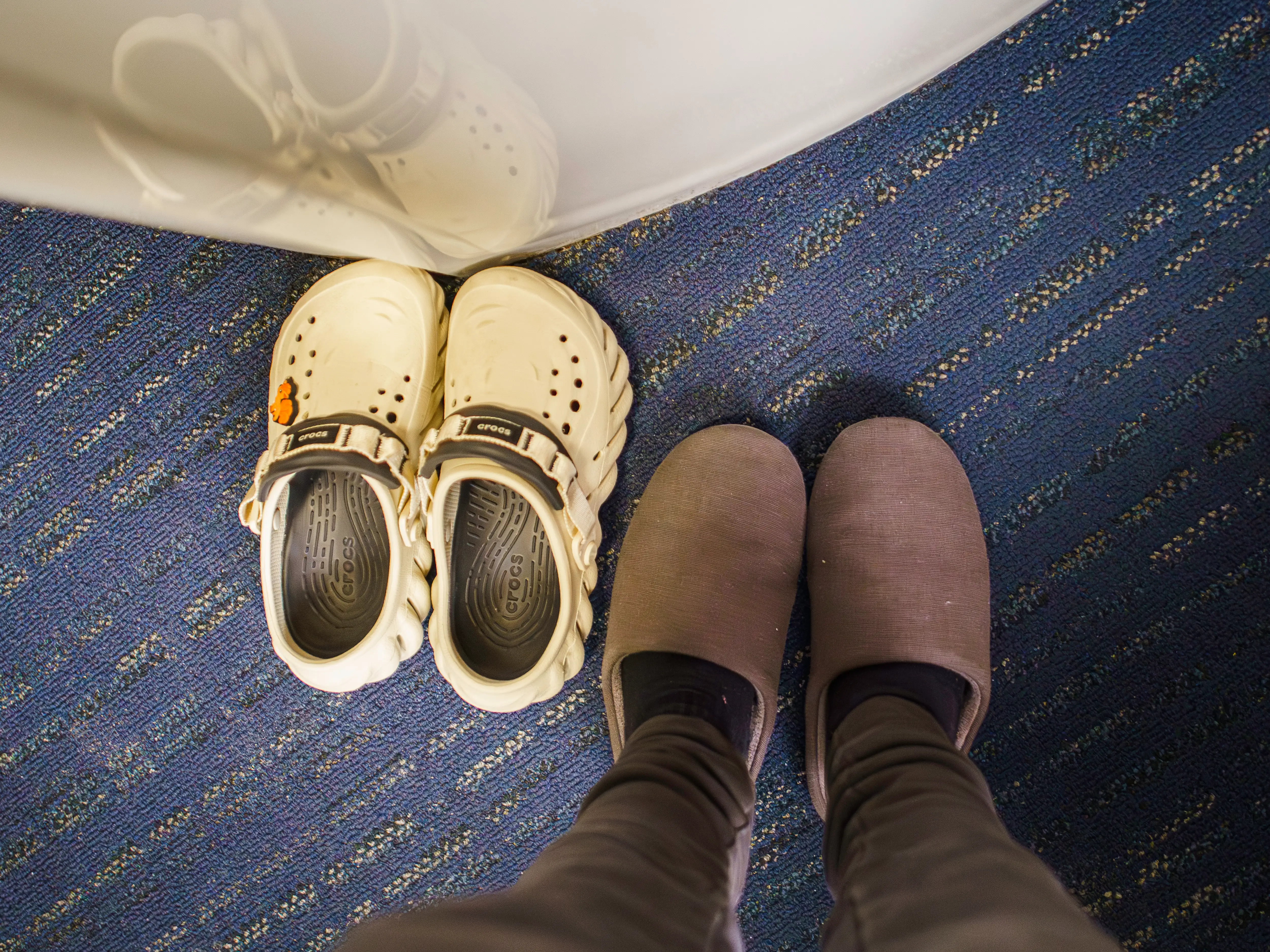 Cream Crocs clogs sit beside slippered feet on a blue patterned carpet near a reflective surface.
