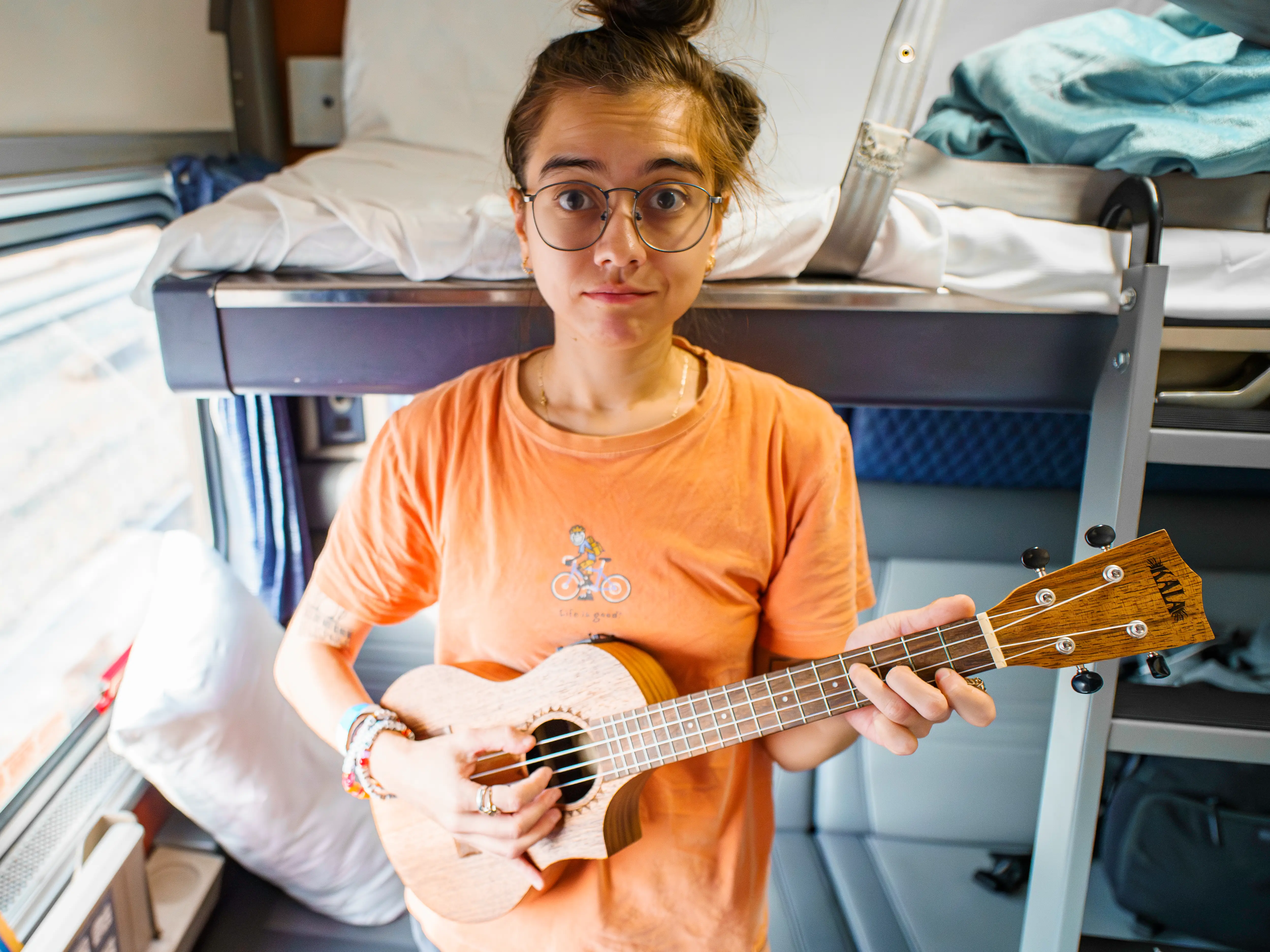 Person with glasses holds a ukulele inside a train sleeper compartment with bunk beds and a window.