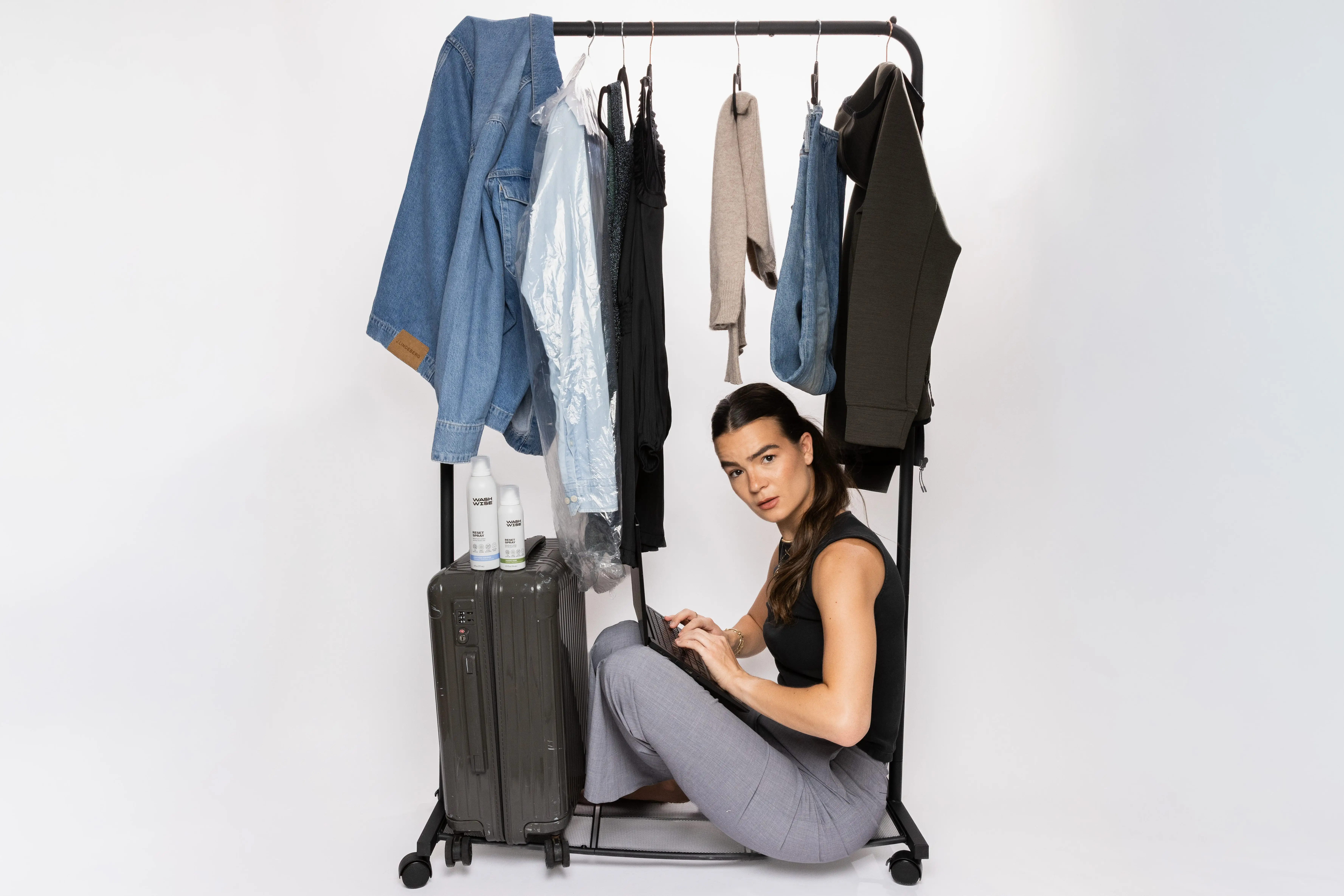 Person seated inside a clothing rack works on a laptop beside hanging clothes, a suitcase, and bottles.