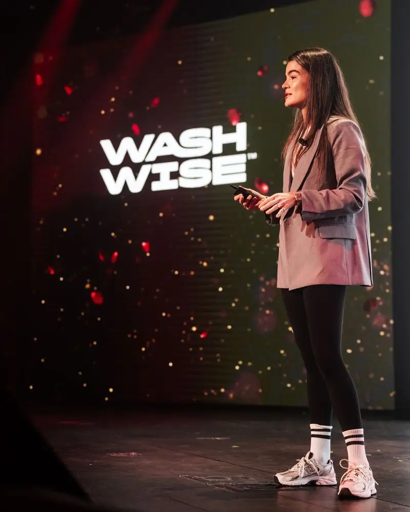 Presenter stands on a stage holding a clicker beside a large screen reading “WASH WISE™.”