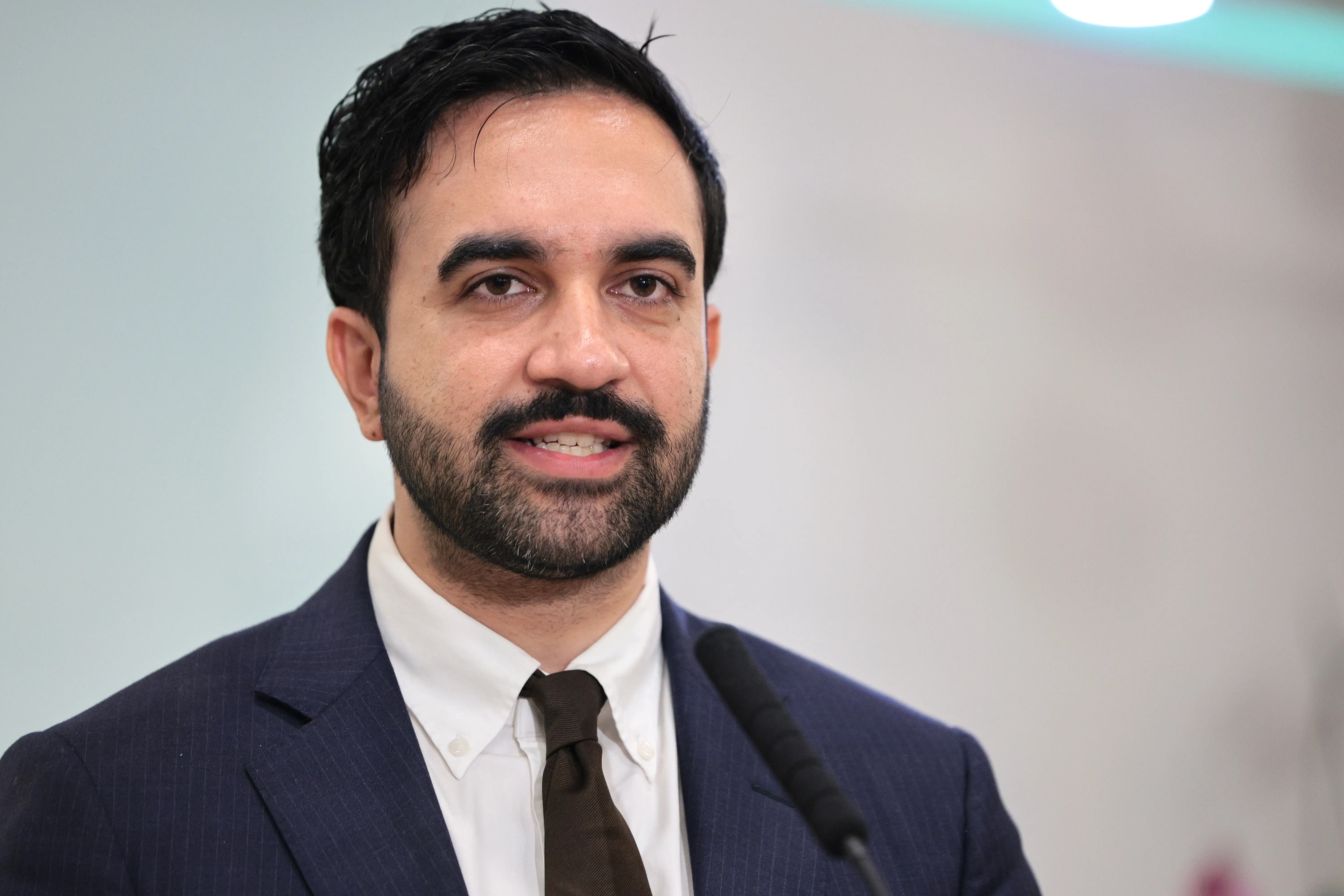 Zohran Mamdani speaks at a microphone while wearing a suit and tie against a blurred indoor background.