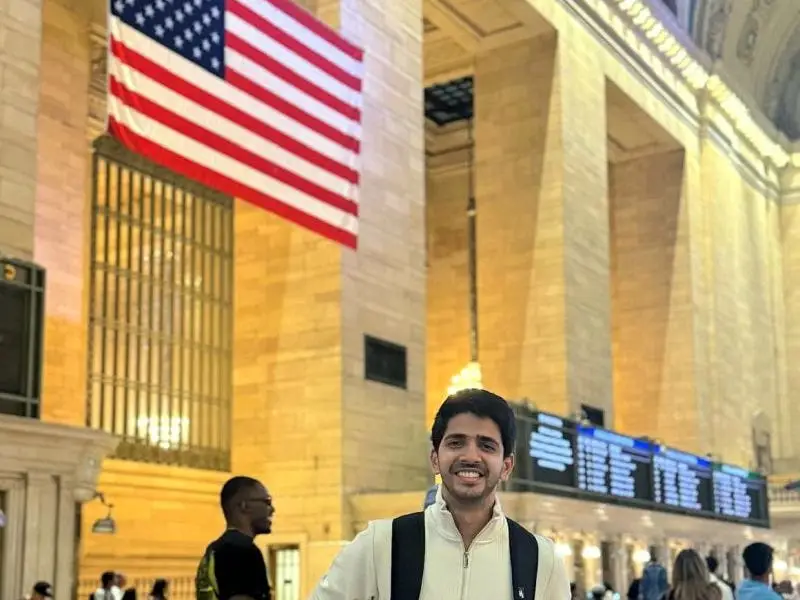 A man wearing a backpack posing below the American flag.