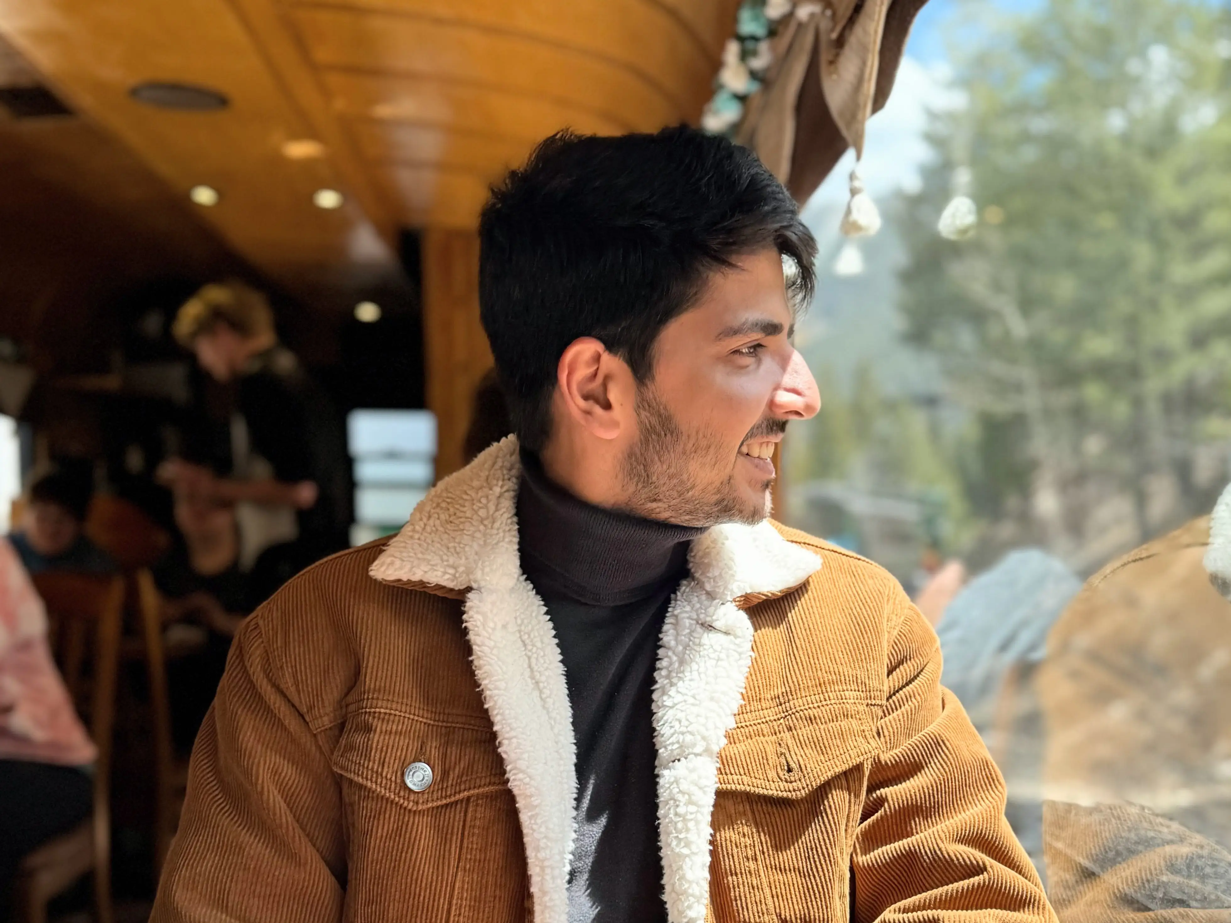 A man wearing a brown courderoy jacket, sitting by the window and looking outside.