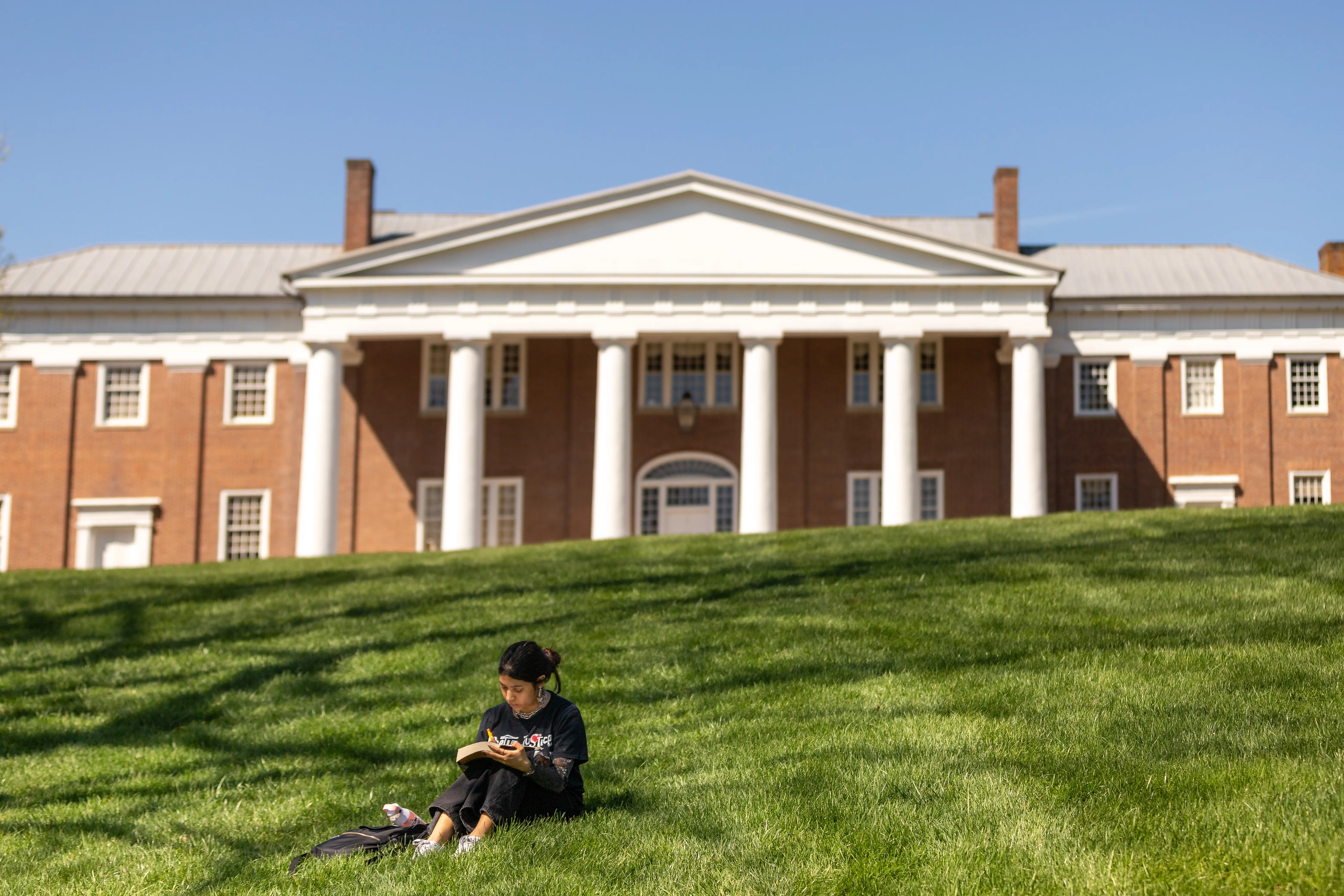 A person sits on a grassy lawn at Centre College, Kentucky with a notebook in front of a large brick building with white columns.