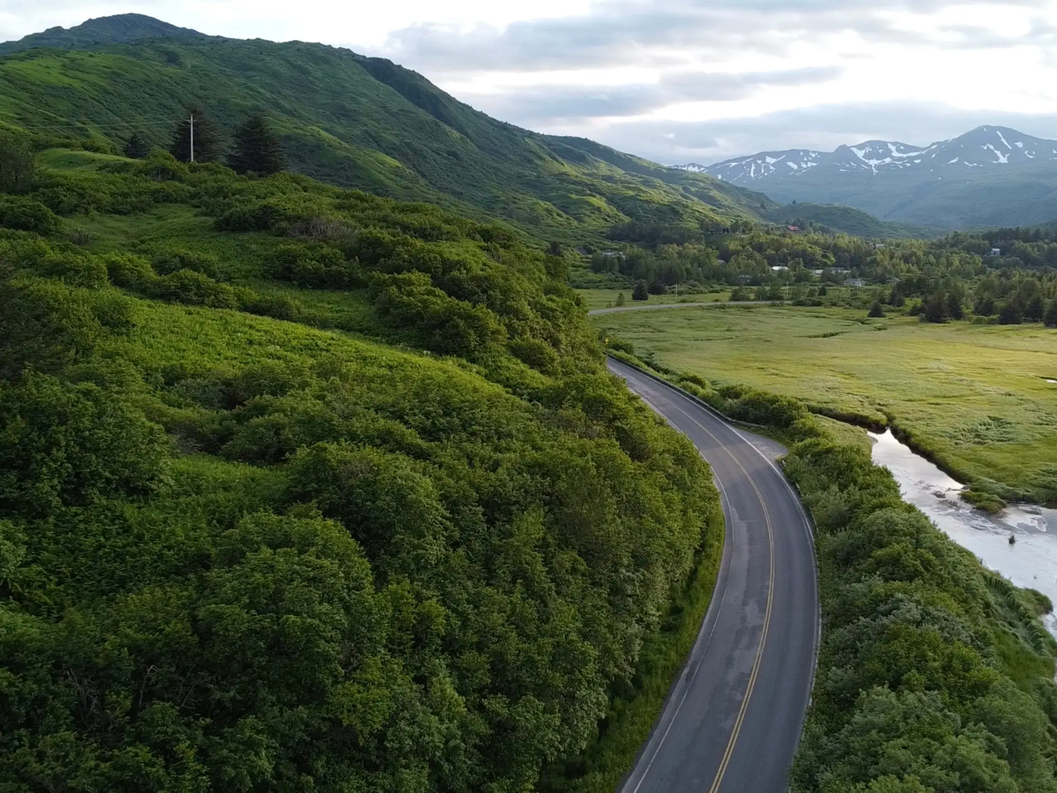 A winding road runs through a green mountain valley beside a narrow river in remote Alaska.