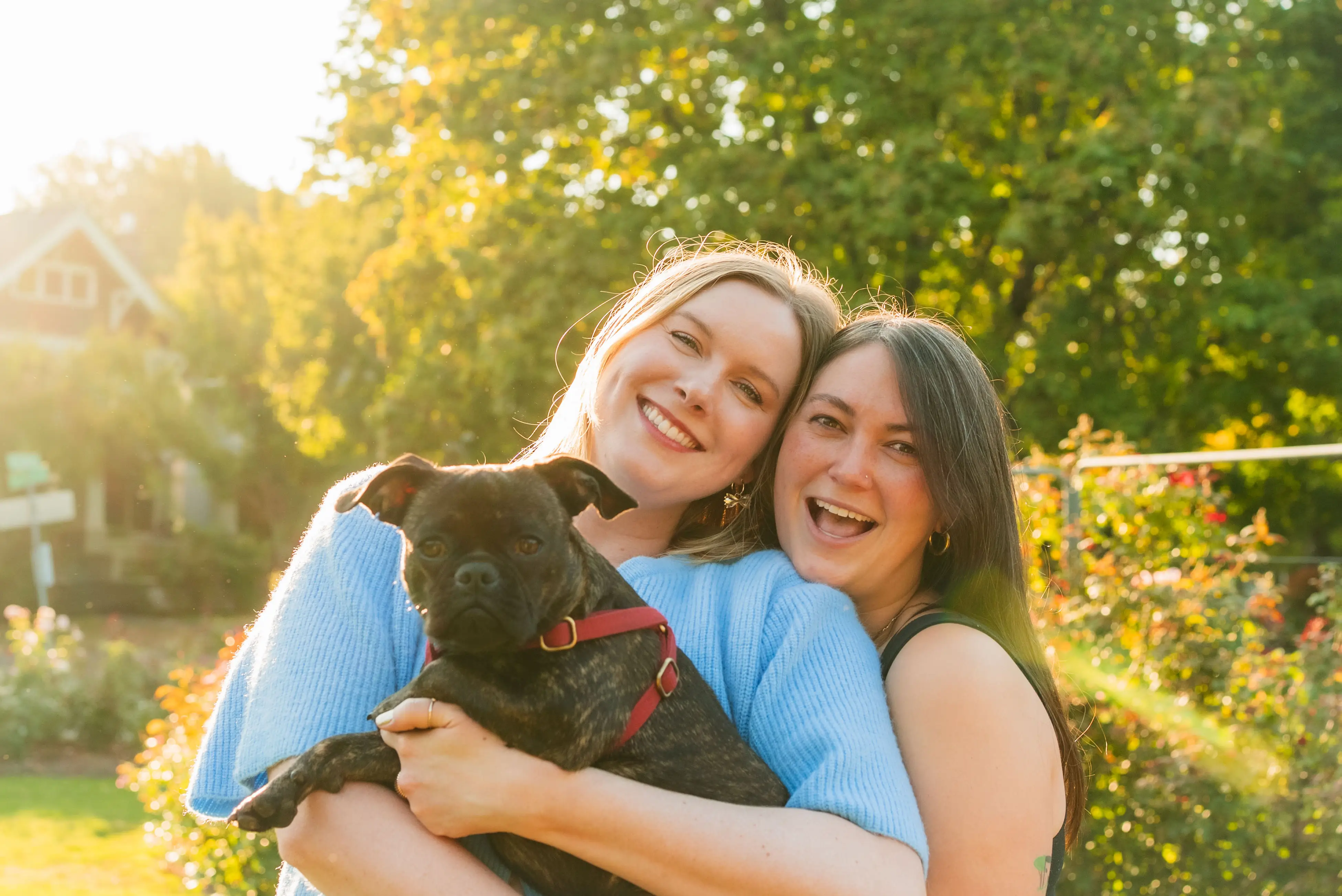 The writer and her partner holding a small dog in front of their house and trees.