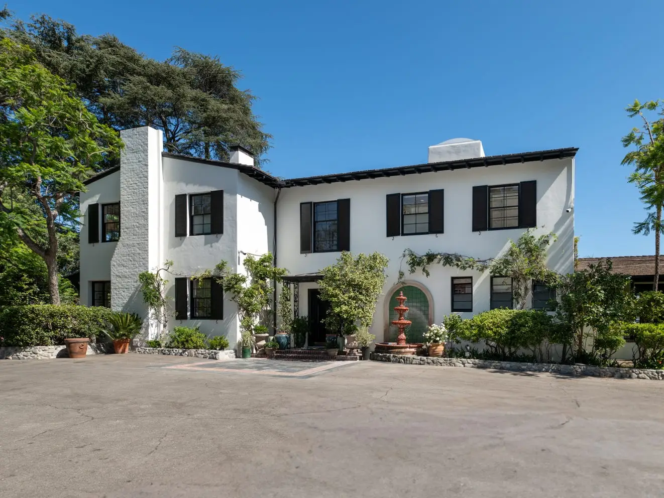 White two-story house with black shutters, a front fountain, landscaped greenery, and a wide driveway under a clear sky.