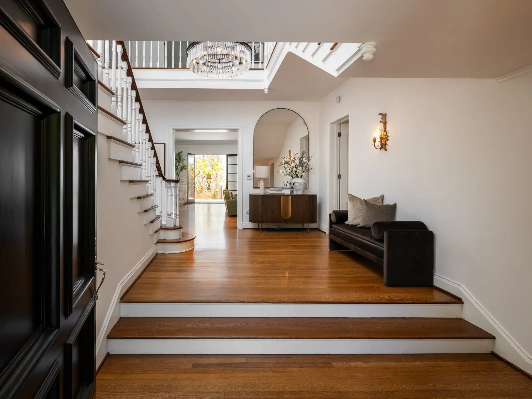 Elegant home entryway with wood floors, staircase, chandelier, console table, arched mirror, and dark upholstered bench.