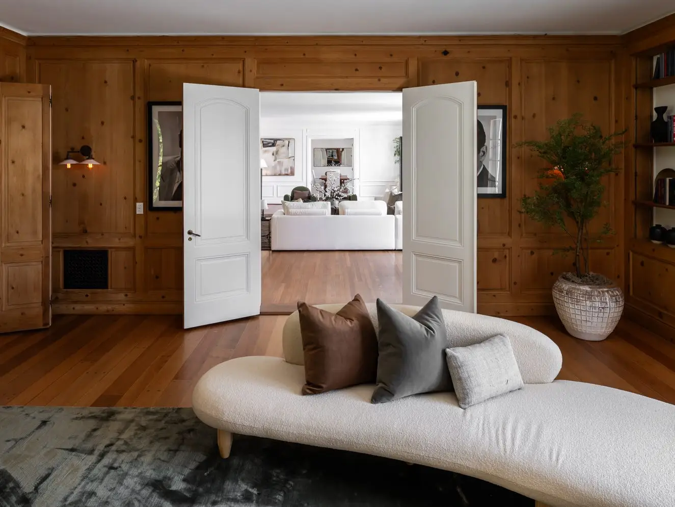 Wood-paneled lounge with a curved white chaise, built-in shelves, open double doors, and a bright adjoining living room.