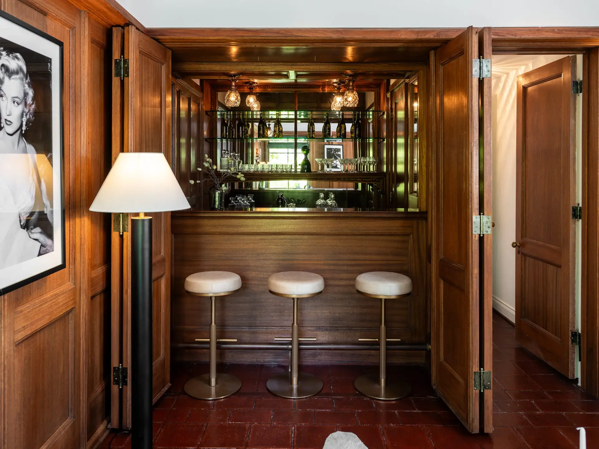 Wood-paneled home bar with three stools, glass shelving, open folding doors, floor lamp, and a framed Marilyn Monroe portrait.