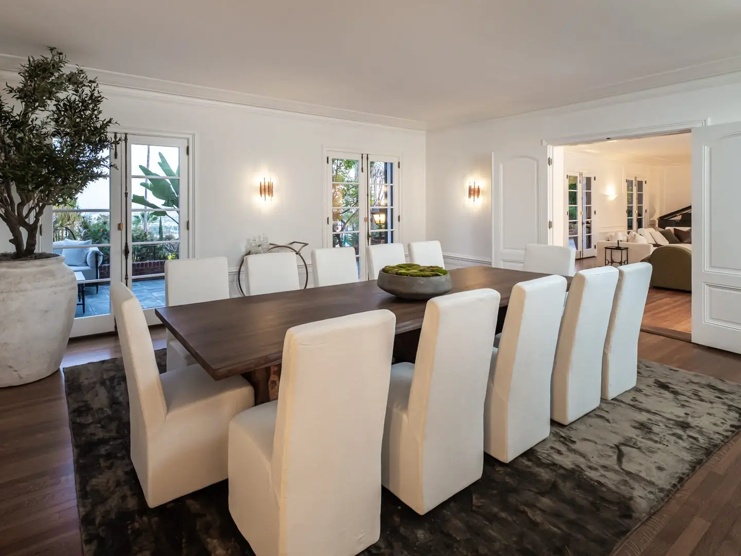 Elegant dining room with a long wooden table, white chairs, potted tree, French doors, and adjoining lounge.