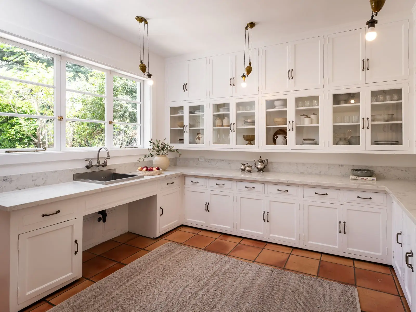 Bright white kitchen with marble counters, glass-front cabinets, pendant bulbs, large windows, and terracotta tile flooring.