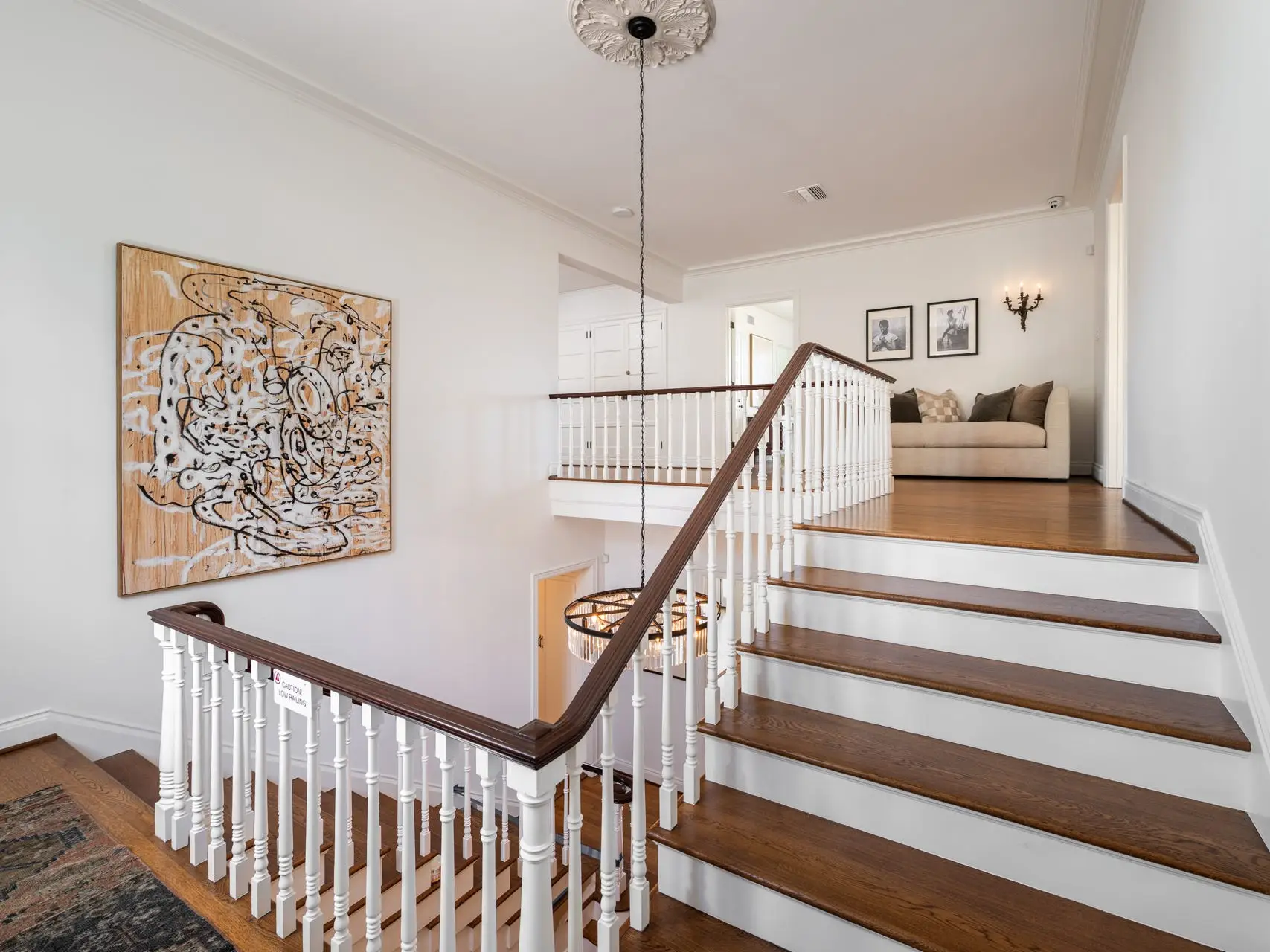Bright interior stair landing with wood steps, white railings, abstract wall art, chandelier, and sofa seating area.