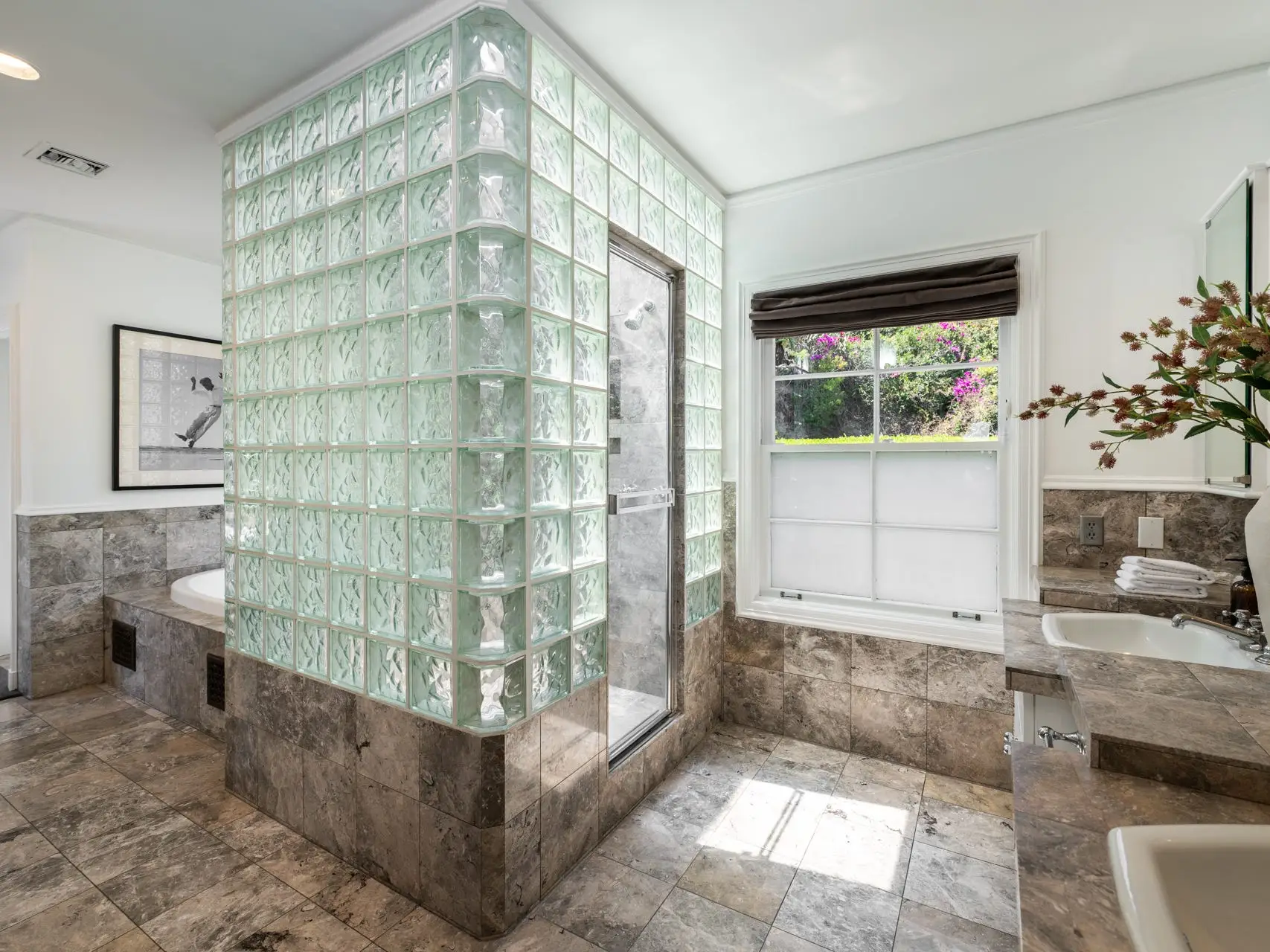 Spacious bathroom with glass-block shower enclosure, stone tile walls and floors, window, bathtub, vanity, and vase of flowers.