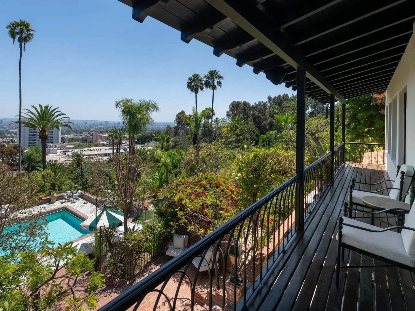 Covered balcony with chairs overlooks a turquoise pool, palm trees, lush hillside landscaping, and a distant city view.