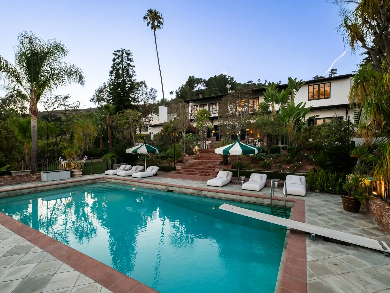 Outdoor swimming pool with lounge chairs, striped umbrellas, palm trees, and a large house at dusk.