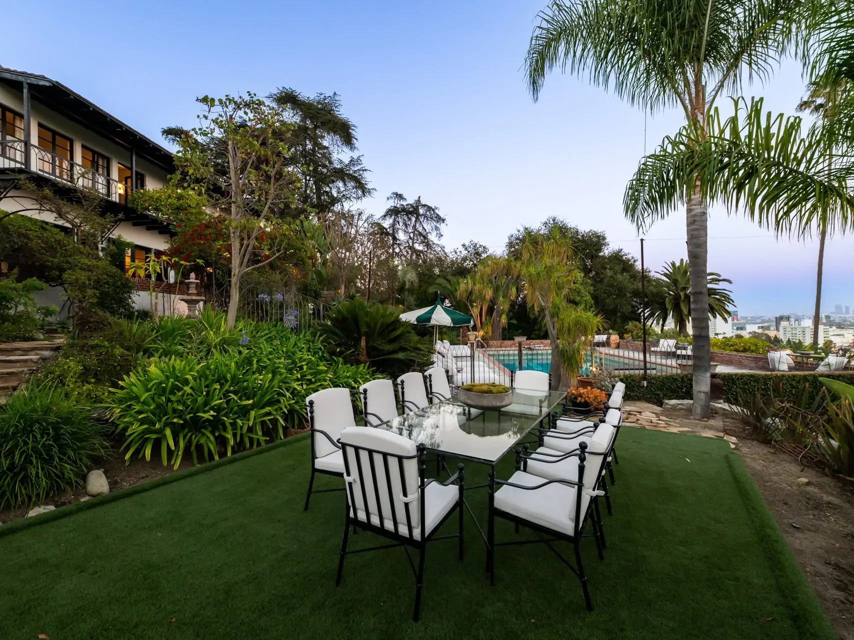 Outdoor dining table with white chairs in a landscaped hillside garden beside a house and pool at dusk.
