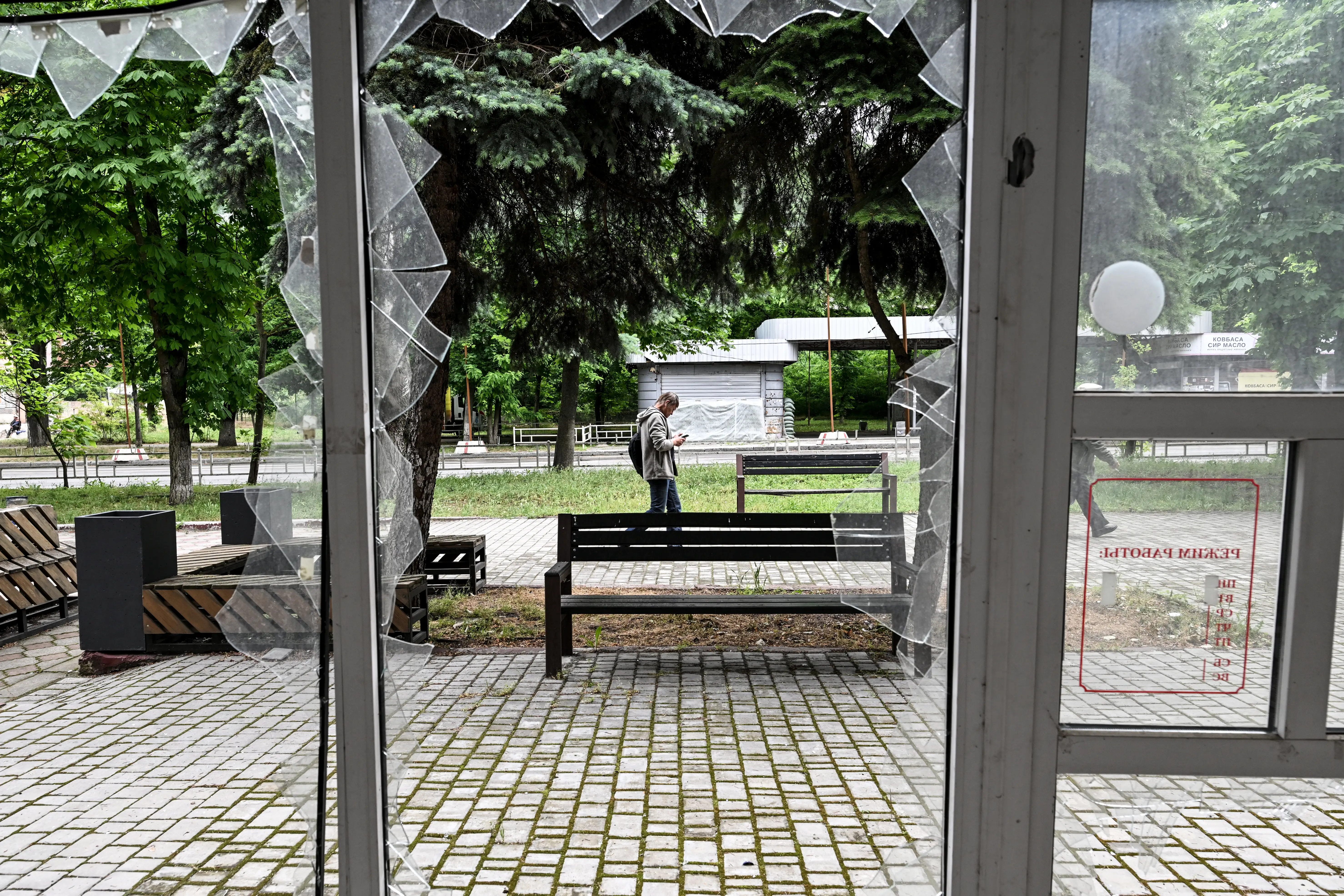 Broken window glass frames a park walkway with benches and a person looking at a phone.