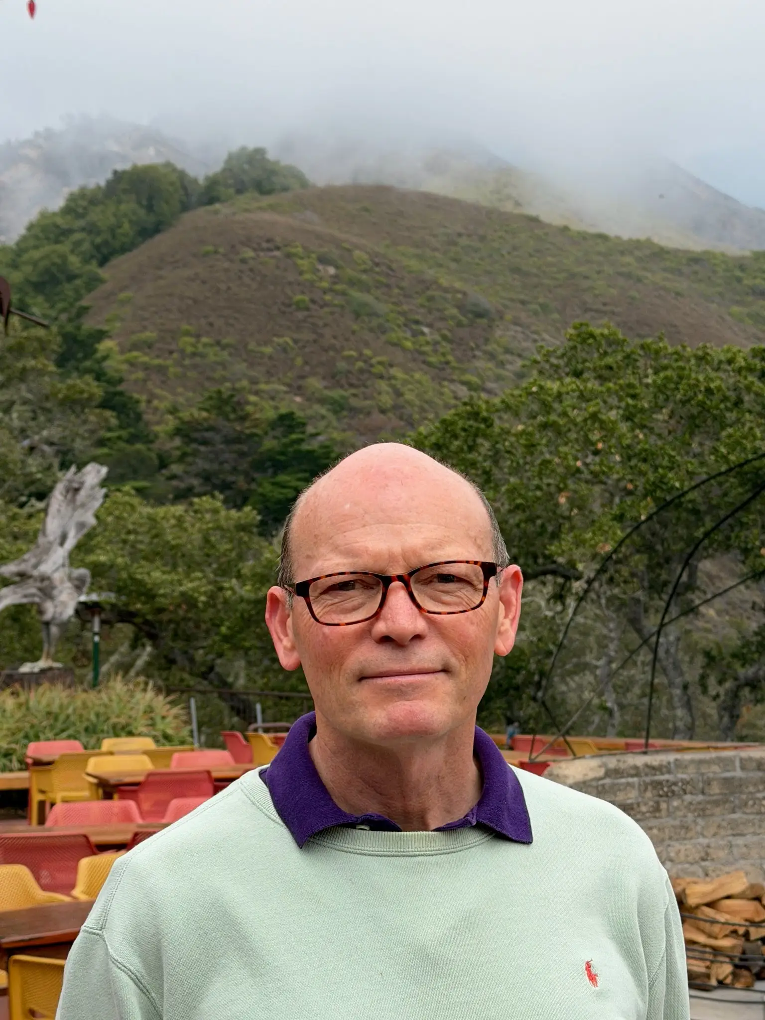 Kirk Gafill wearing glasses stands on an outdoor terrace with colorful chairs and misty green hills behind.